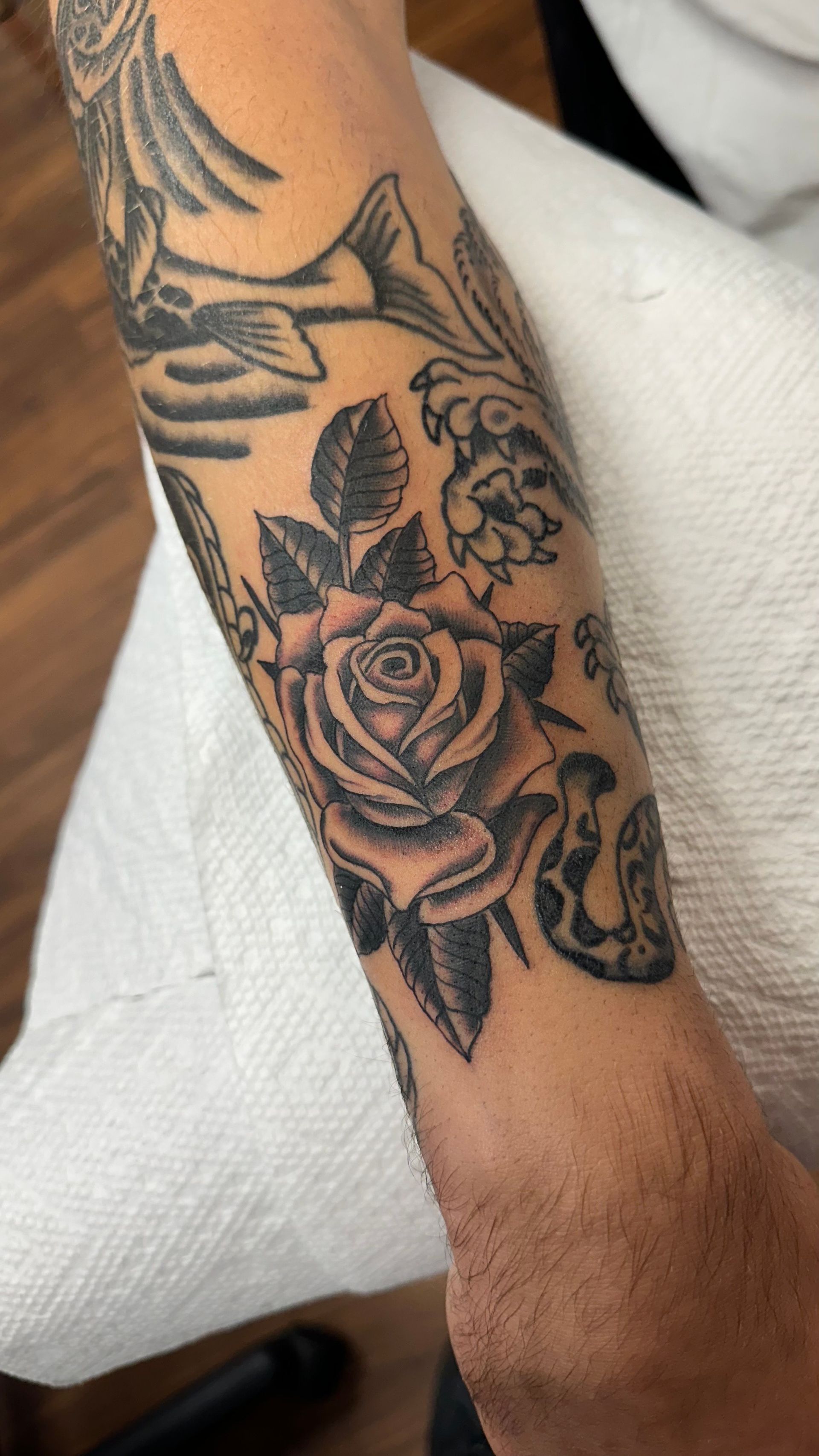 Forearm tattoo of a black and gray rose with leaves, surrounded by other traditional style tattoos.