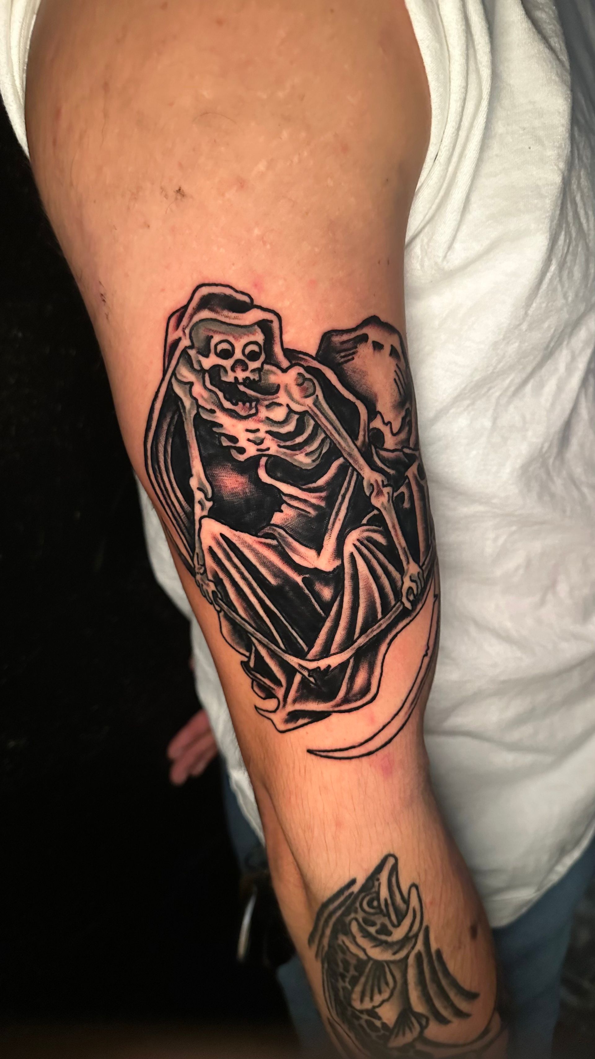 Tattoo of a skeleton with a scythe on an arm. Black and white, sitting on an arch.