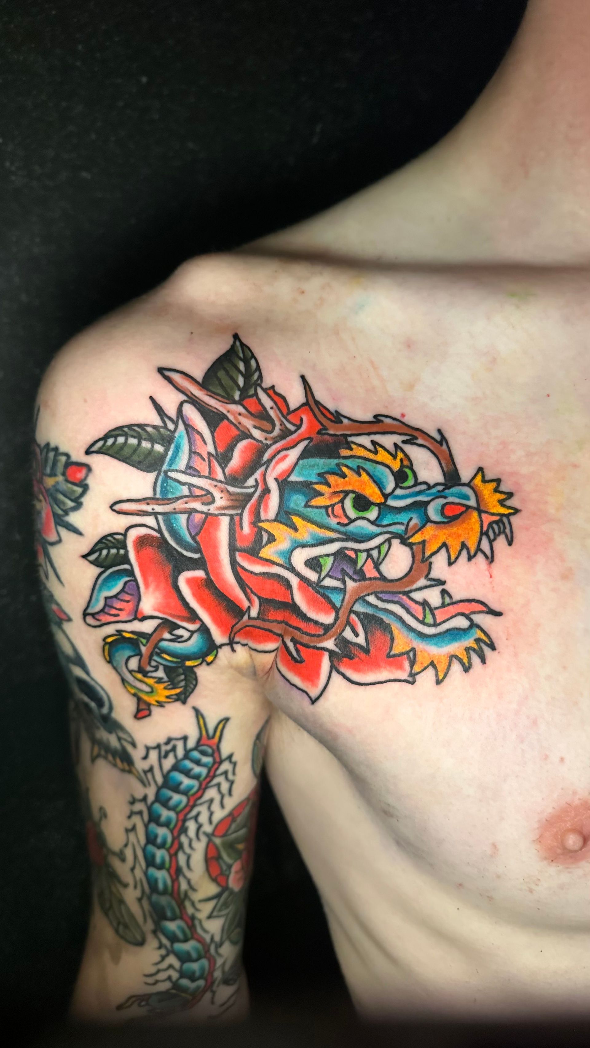 Dragon's head emerging from a red rose tattoo on a person's shoulder and chest; multi-colored ink.