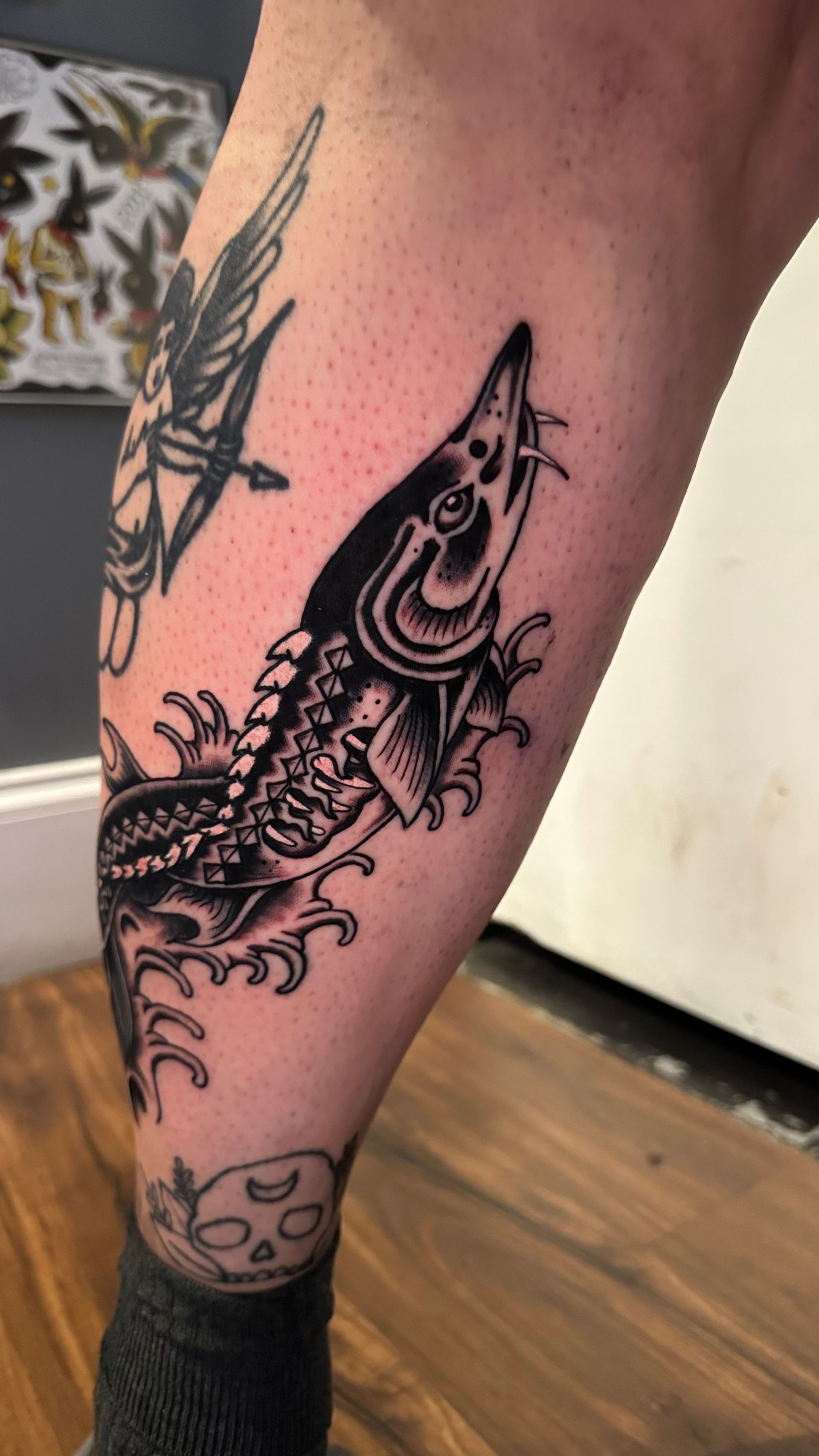 Black and white tattoo of a fish on a leg. The fish has spiky scales, open mouth, and is surrounded by water-like details.