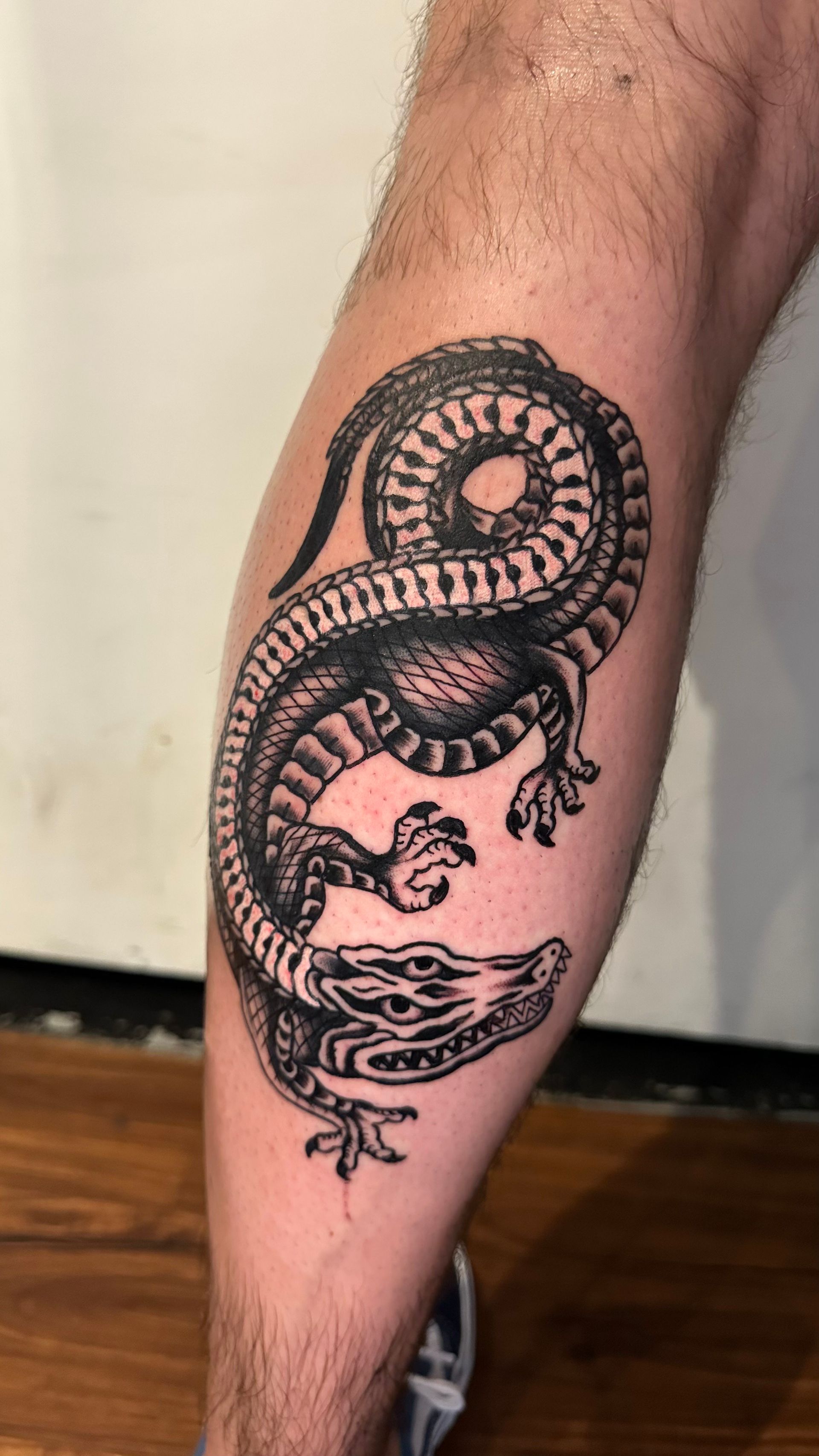 Black ink tattoo of an alligator on a leg. The alligator is coiled with detailed scales and claws.