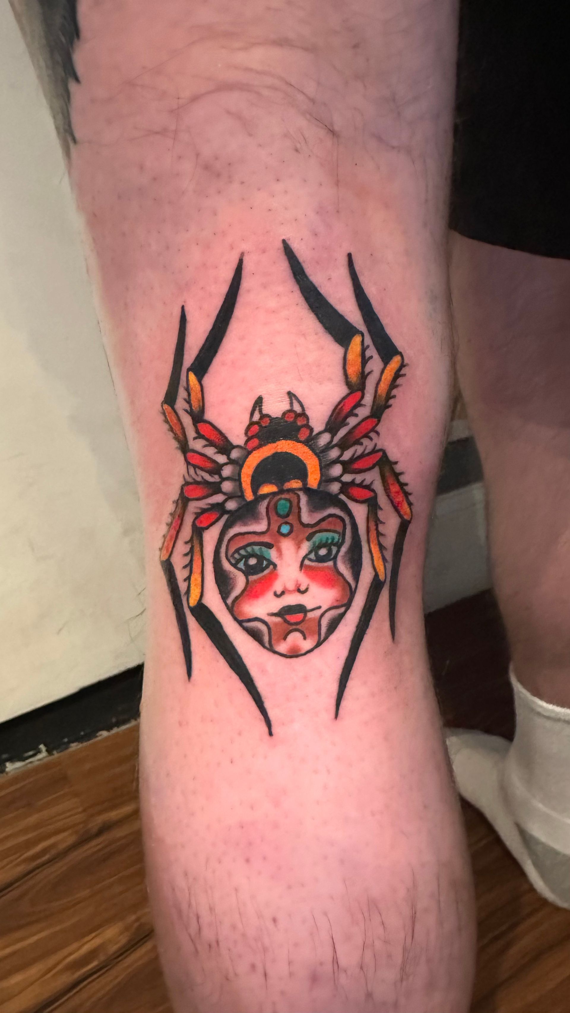 Tattoo of a spider with a face on a person's knee. Red, yellow, and black colors. Traditional style.