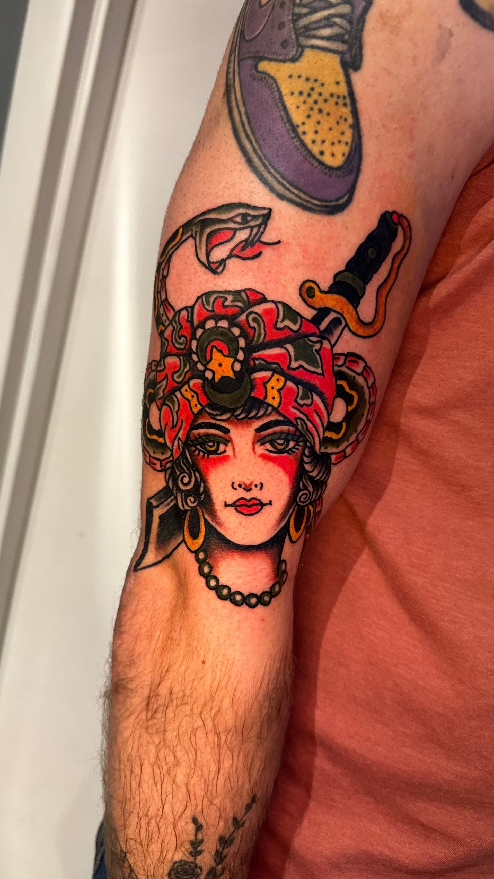 Tattoo of Medusa head with snakes, dagger, and ornate headdress on an arm; red, black, and gold colors.