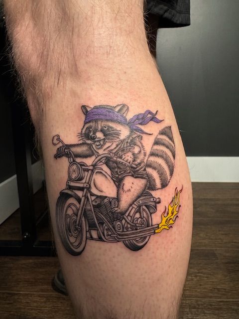 A tattoo of a raccoon riding a motorcycle on a leg