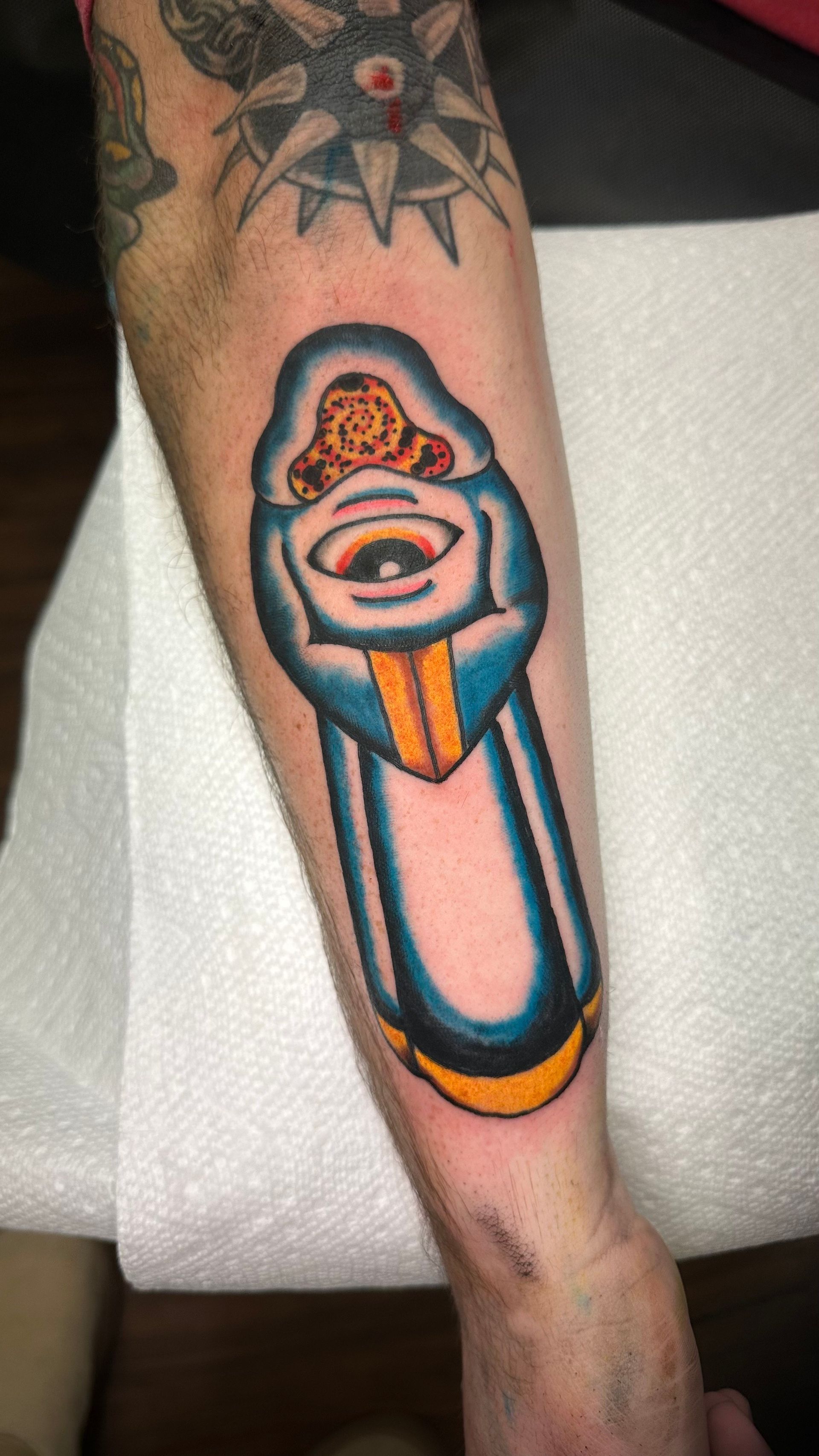 Tattoo of a hooded figure holding a centered eye, with blue, yellow and brown colors on an arm.