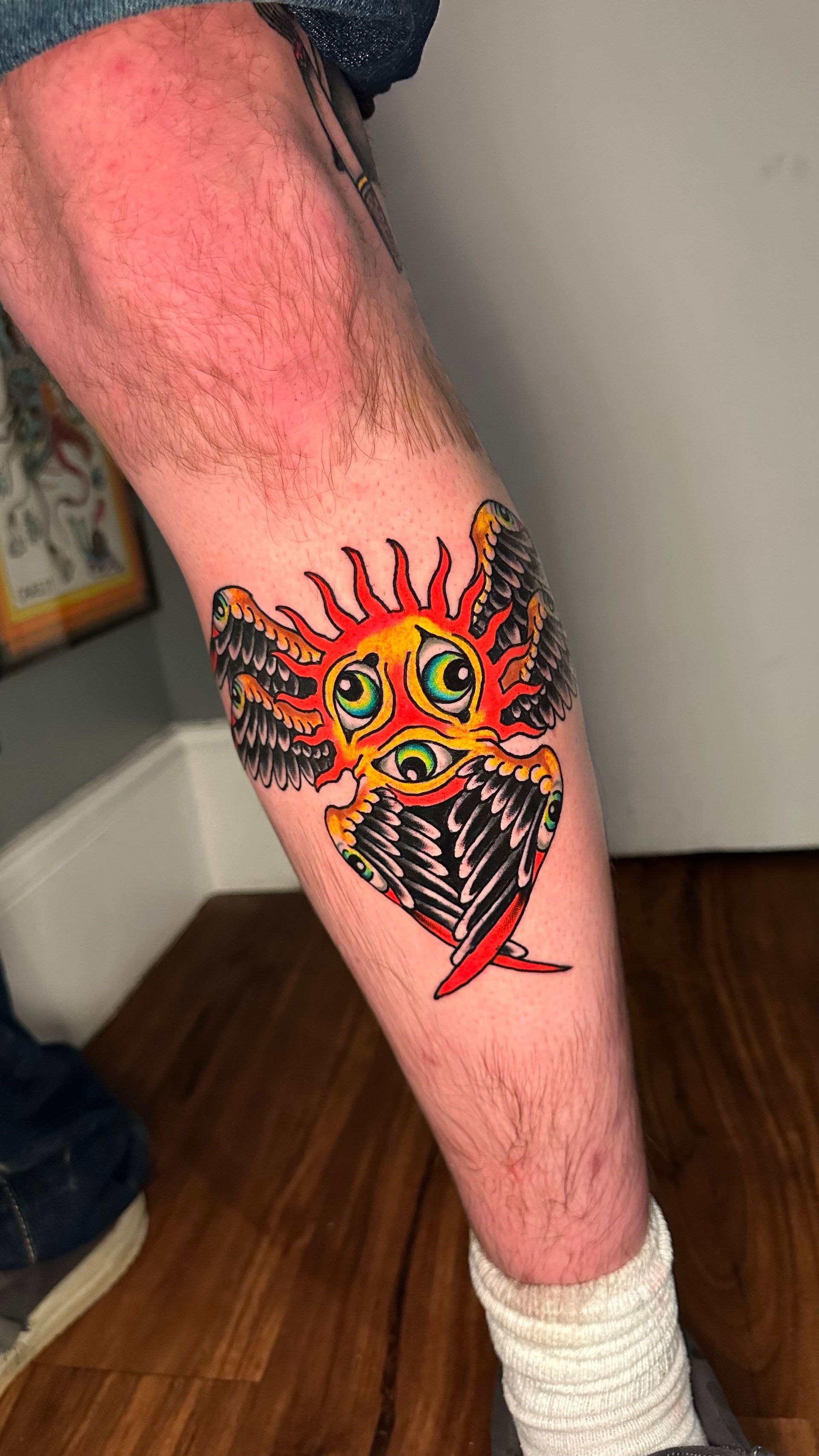 Tattoo of a whimsical sun with wings and three eyes, on a person's calf.