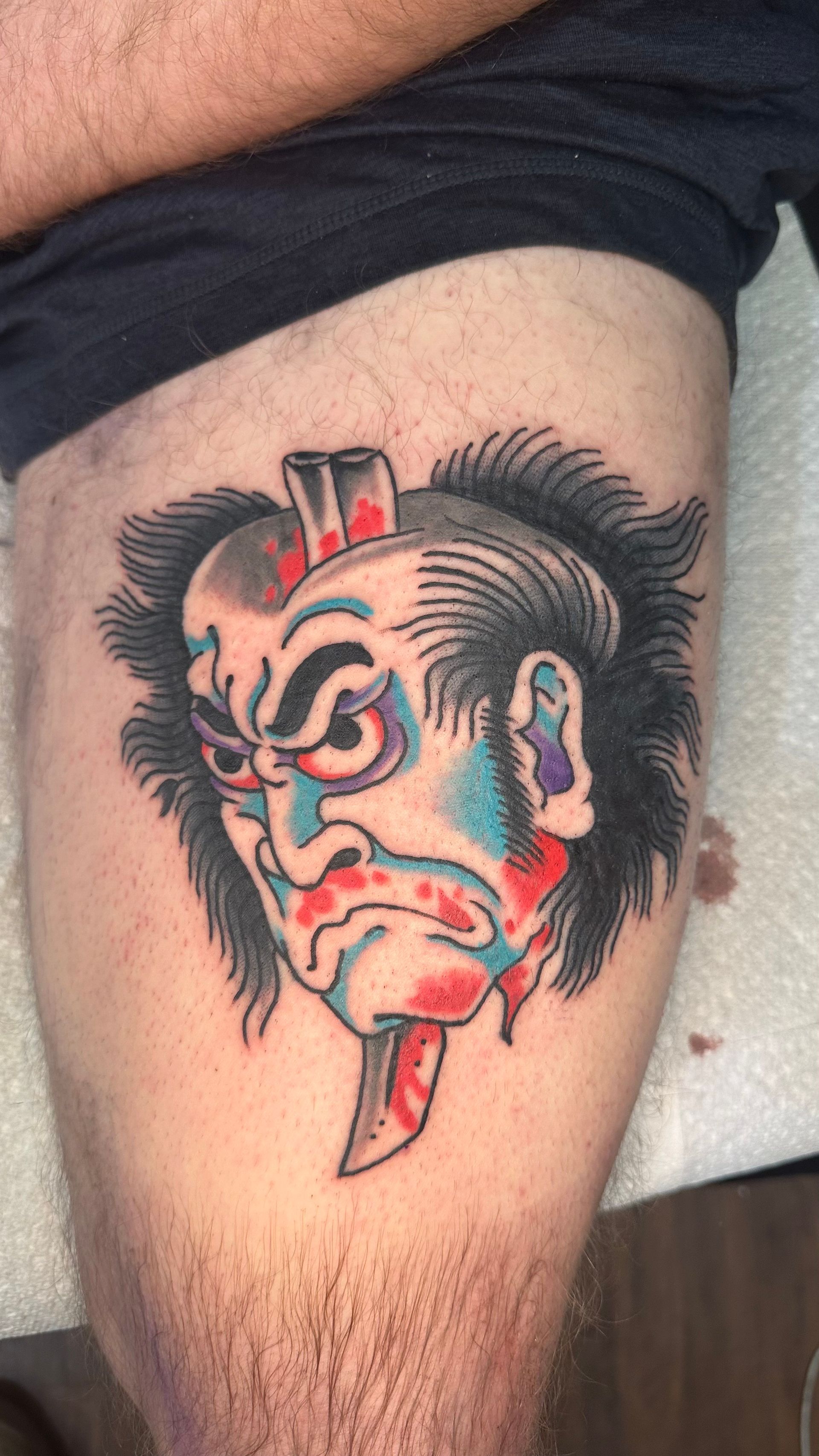 Tattoo of a stylized, bloody Japanese demon mask on a leg, with blue, red, and black ink.
