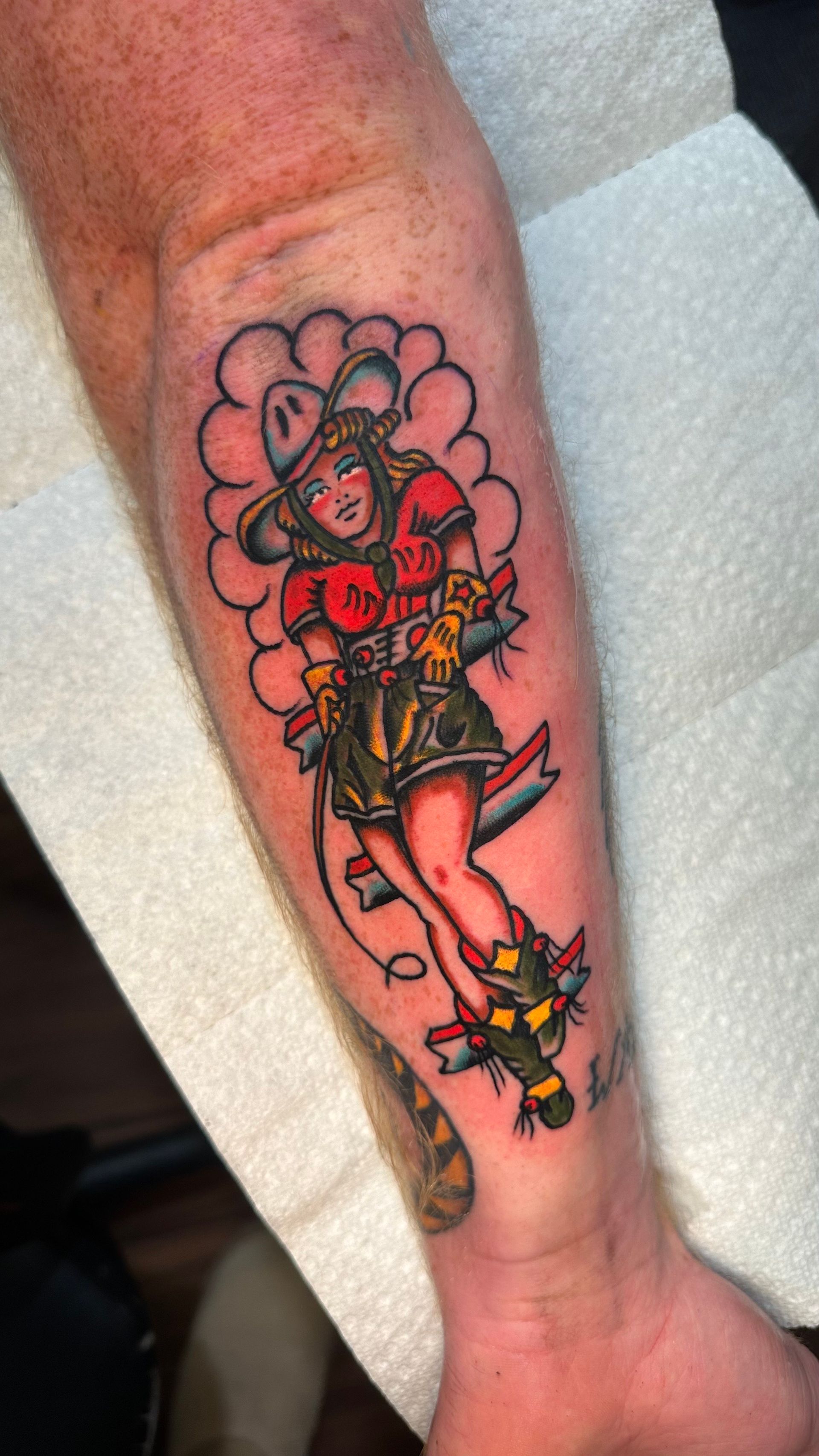 Tattoo of a woman in cowgirl attire, surrounded by a floral design, on a forearm.