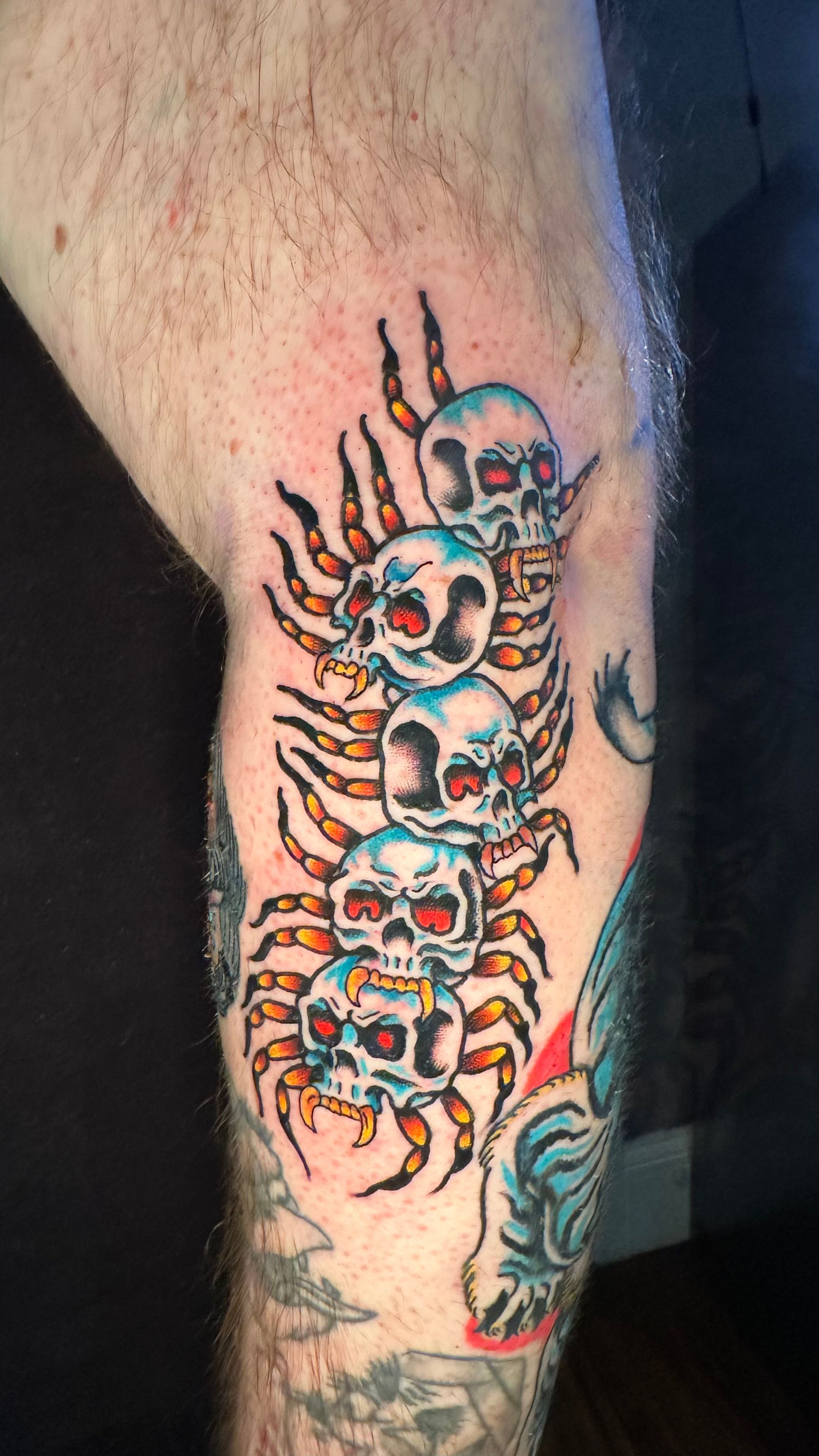 Tattoo of stacked skulls, with spider-like legs, in red, orange, and blue on a leg.