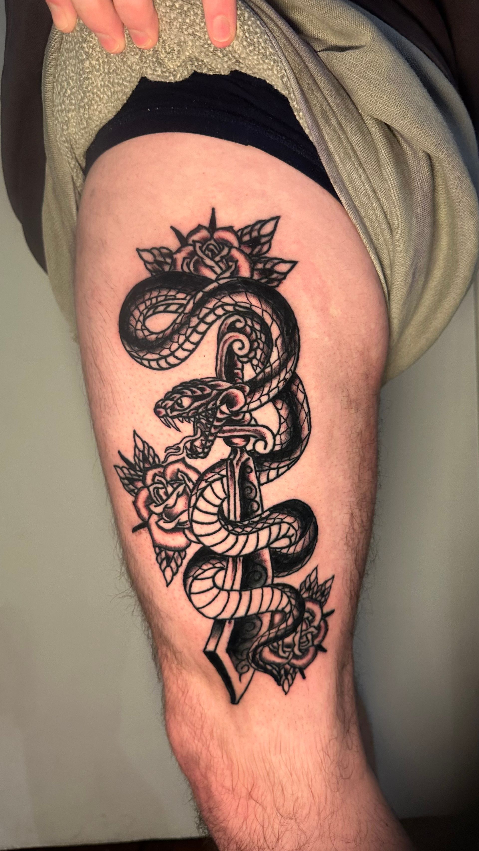 Black and white snake tattoo on thigh, entwined around a blade, with roses and triangular shapes.