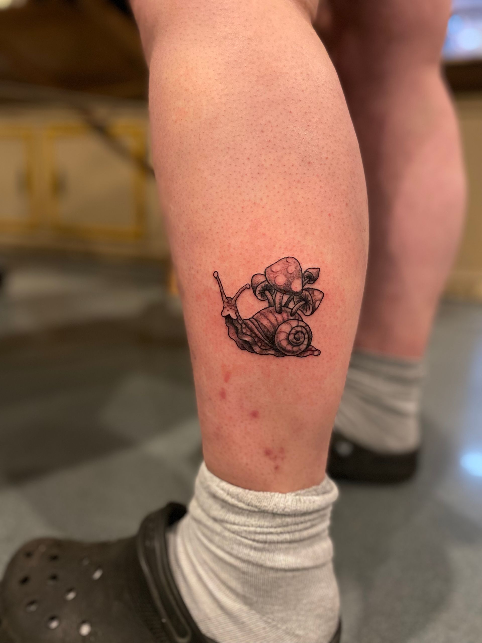 Small black snail and mushroom tattoo on the leg. A unique design by one of the best tattoo parlors in North Carolina.