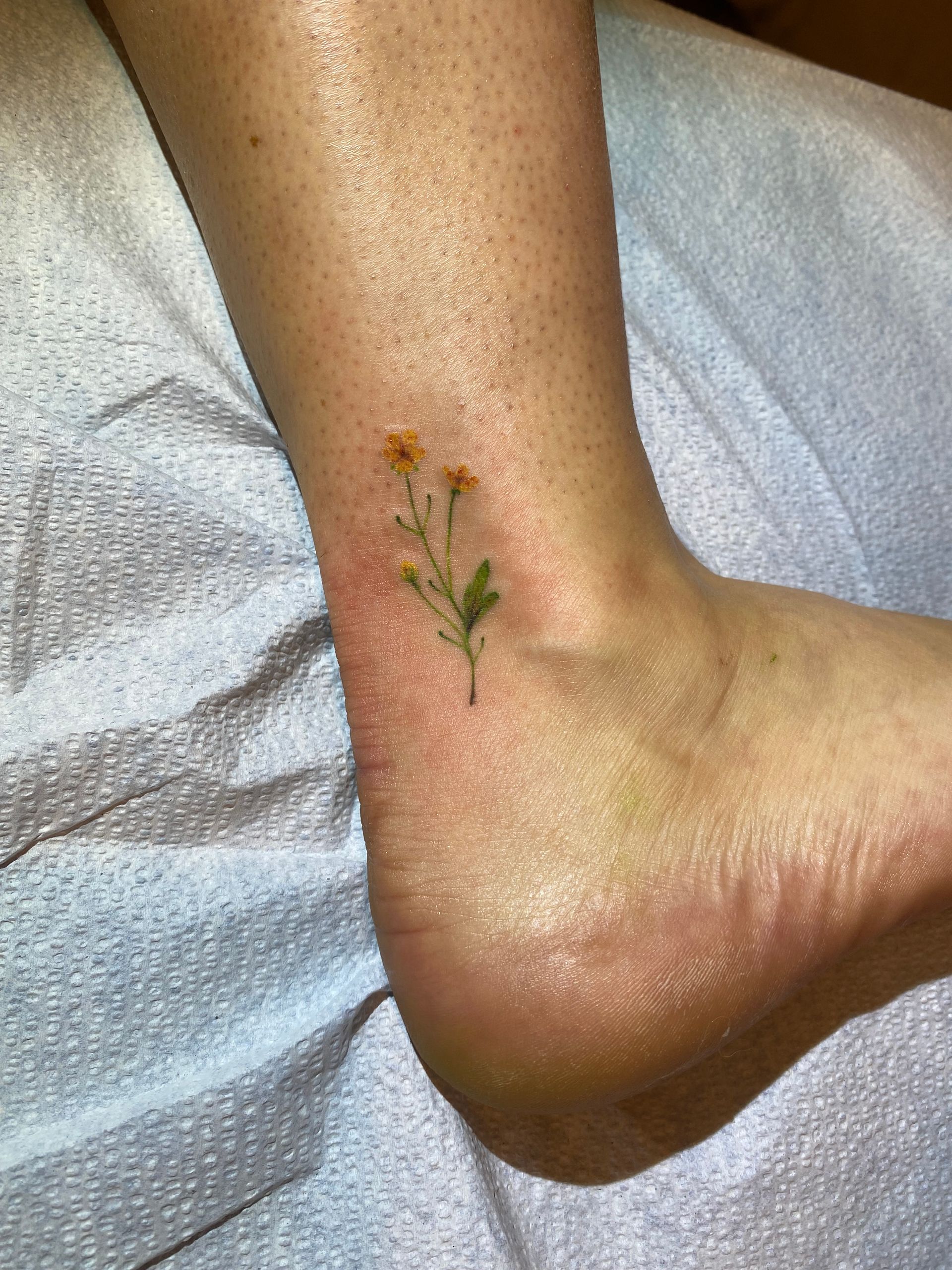 Fine line yellow flower ankle tattoo, nature-inspired design in Raleigh.