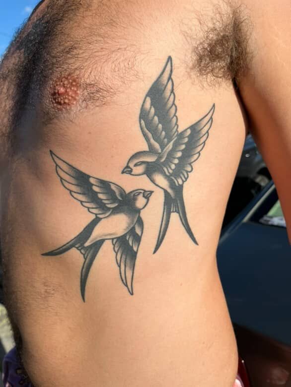 Tattoo of two birds in flight on a person's side, gray and black shading.