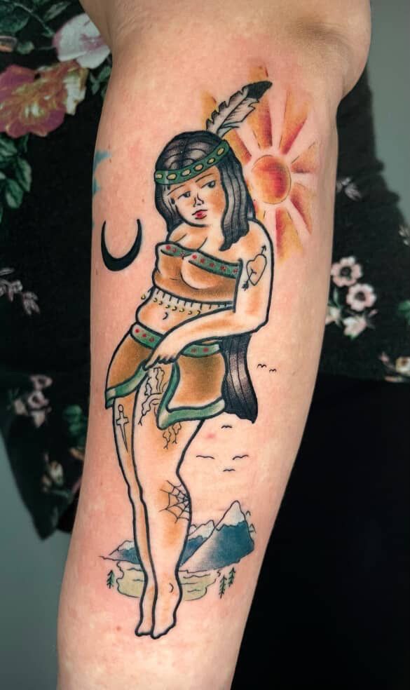 Tattoo of a Native American woman, sun, crescent moon, and mountains on an arm. Brown, black, yellow, and blue colors.