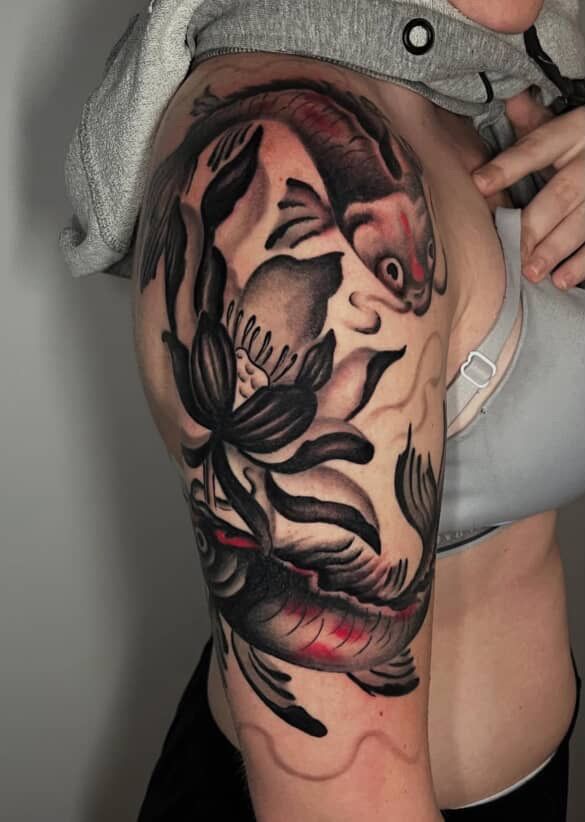 Koi fish tattoo on a person's upper arm, featuring dark fish, a lotus flower, and grey and red accents.