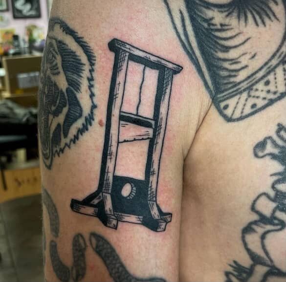 Black and gray guillotine tattoo on arm.