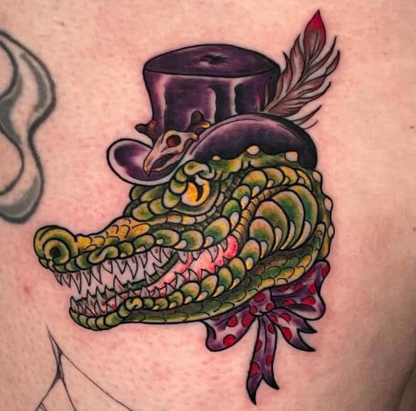 Tattoo of a green alligator head wearing a purple top hat, red feather, and polka dot bow tie.