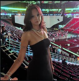 Woman in a black dress posing in a stadium, with red seats and arena lights in the background