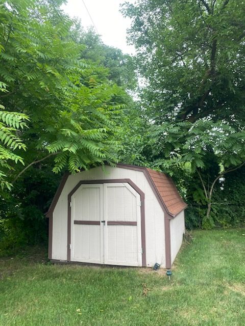 A white shed with a brown trim is surrounded by trees in a yard.