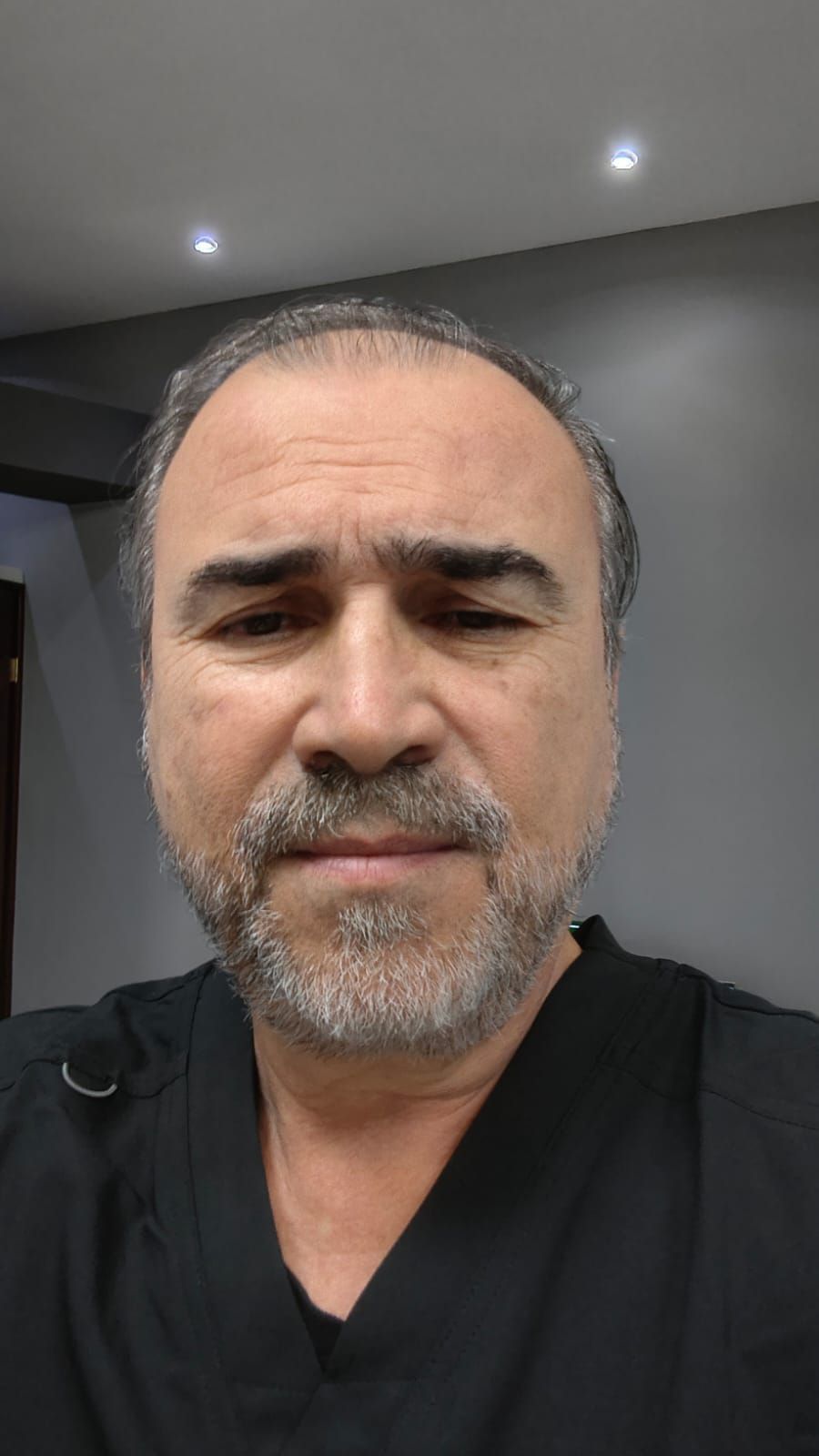 Una persona con barba, vestida con una camisa negra, mira directamente a la cámara en una habitación con iluminación empotrada en el techo.