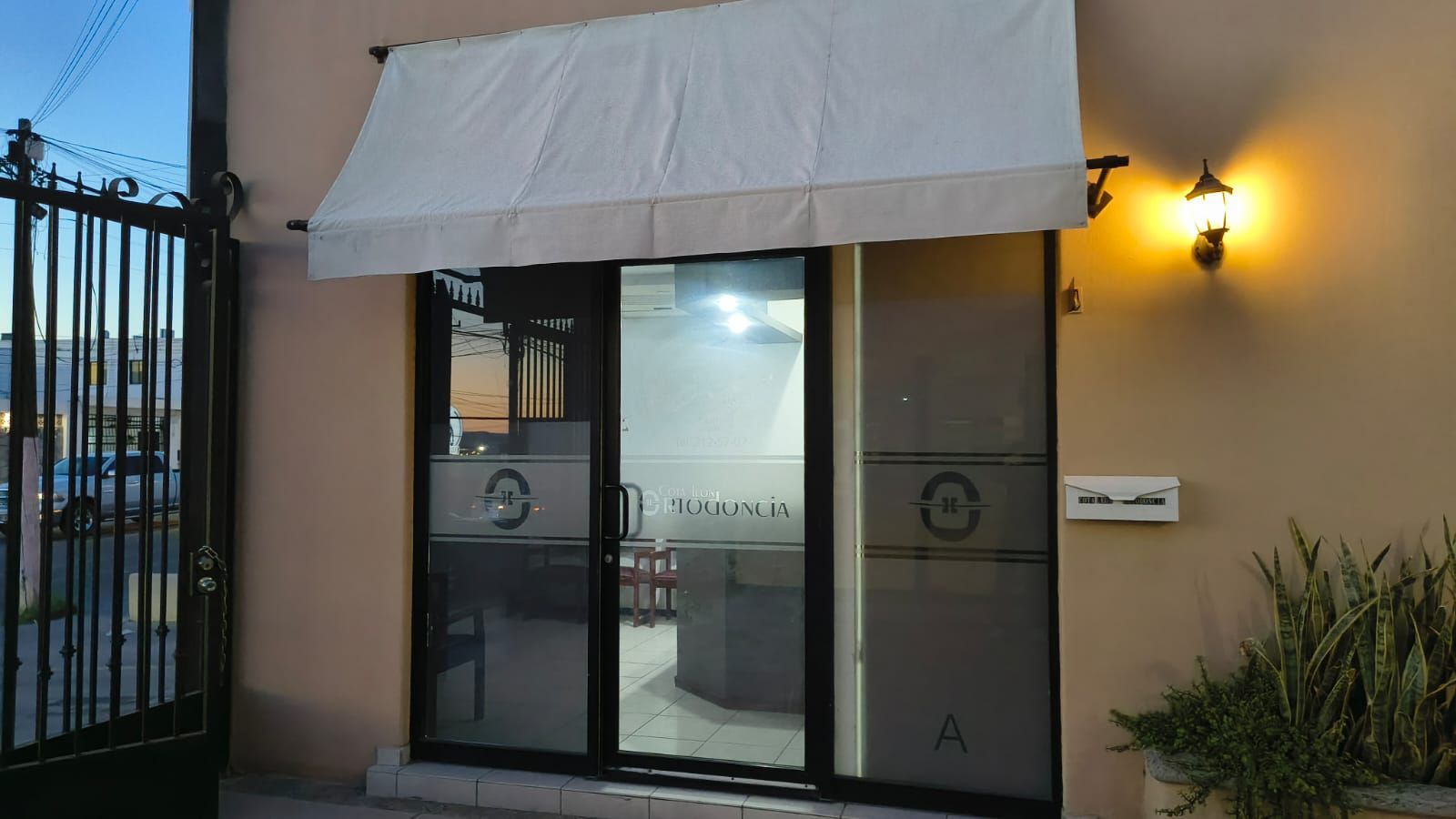 Fachada exterior de color tostado, toldo de tela beige, puertas dobles de cristal y una pequeña luz en el porche.