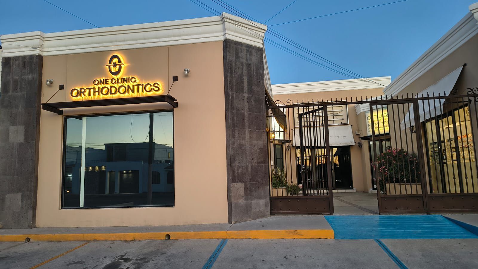 Vista exterior de Gertz Orthodontics, un edificio de color beige con un letrero iluminado, entrada con verja y marcadores azules para los espacios de estacionamiento.
