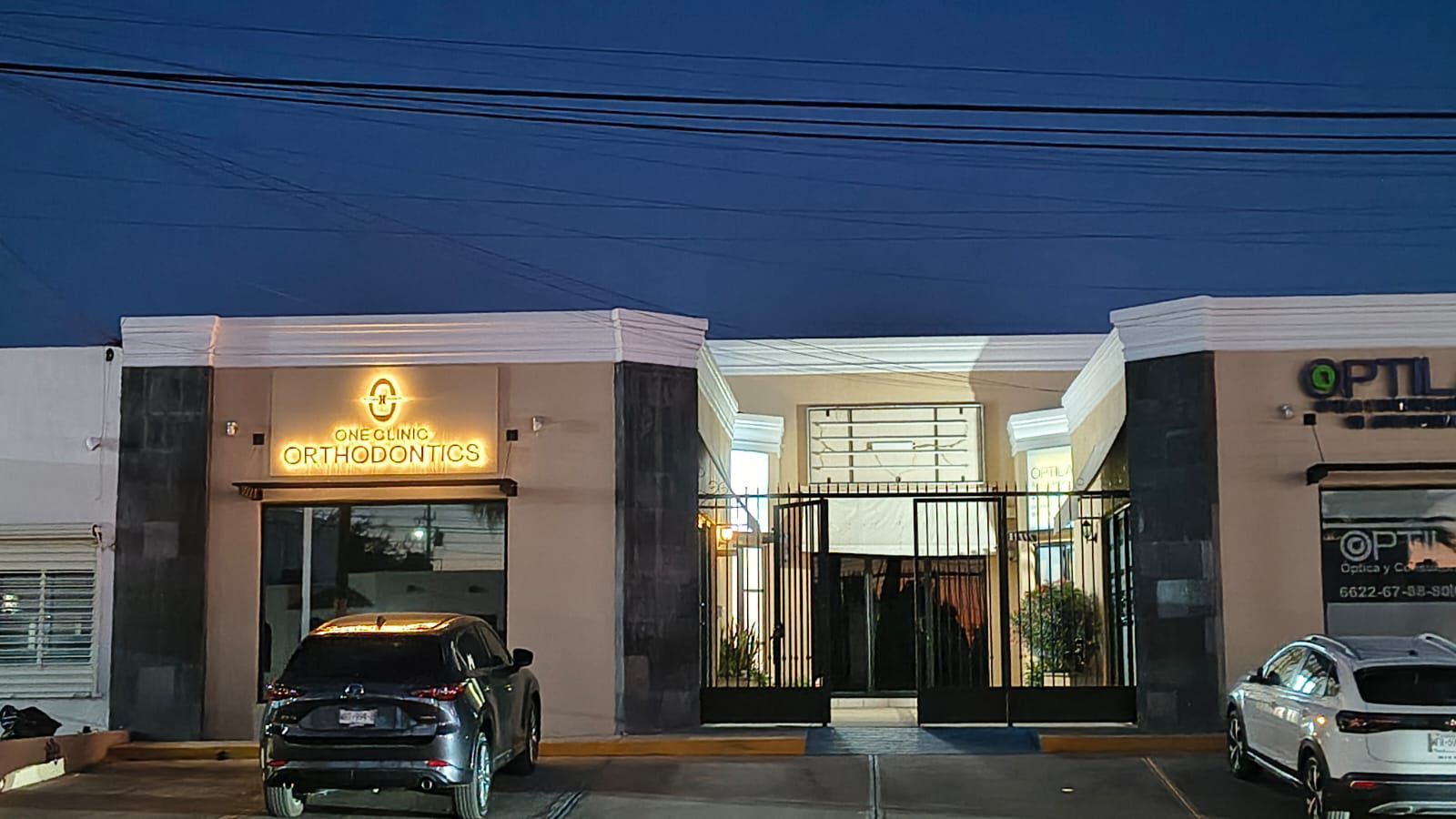 Un edificio comercial de una sola planta al anochecer, con un letrero iluminado, una verja central de hierro y coches aparcados delante.