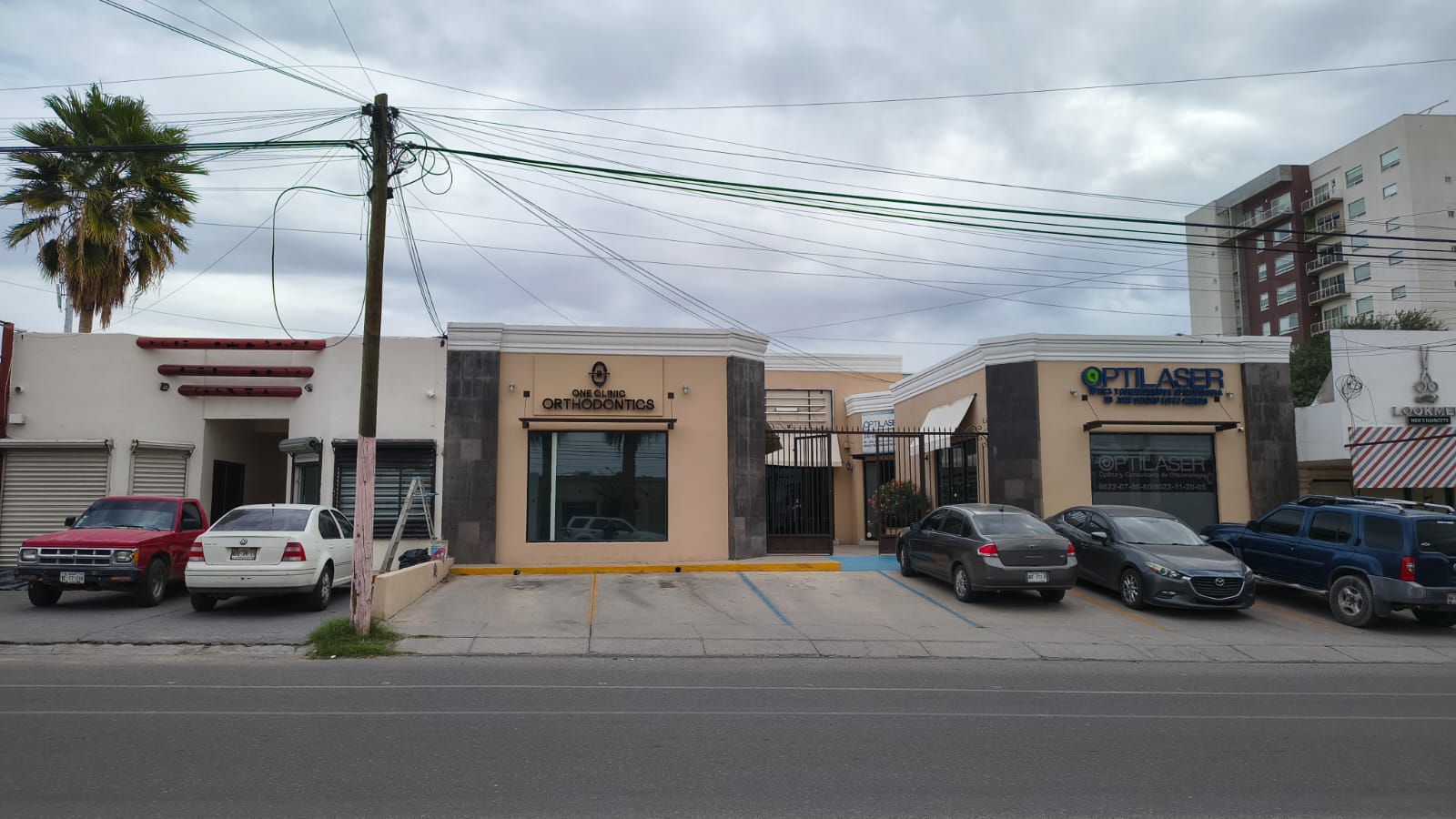 Una hilera de edificios comerciales de color beige y blanco con escaparates, coches aparcados y una palmera bajo un cielo nublado.