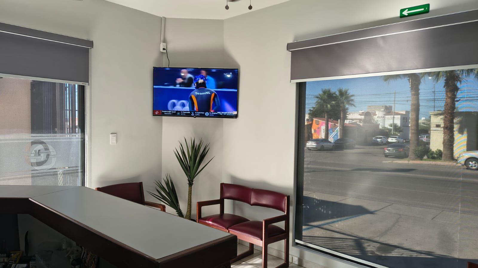 Una sala de espera con un televisor montado en la pared, una pequeña planta en la esquina y dos sillas rojas junto a una ventana con vistas a la calle.
