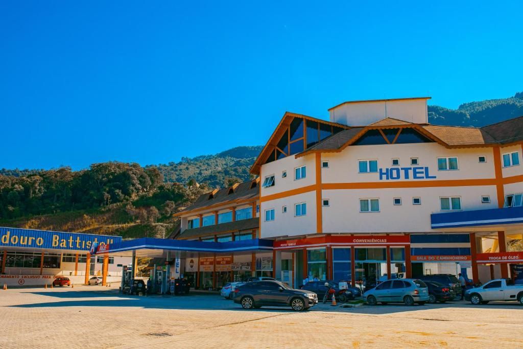 Hotel com posto de gasolina, carros estacionados do lado de fora, montanhas ao fundo, céu azul claro.