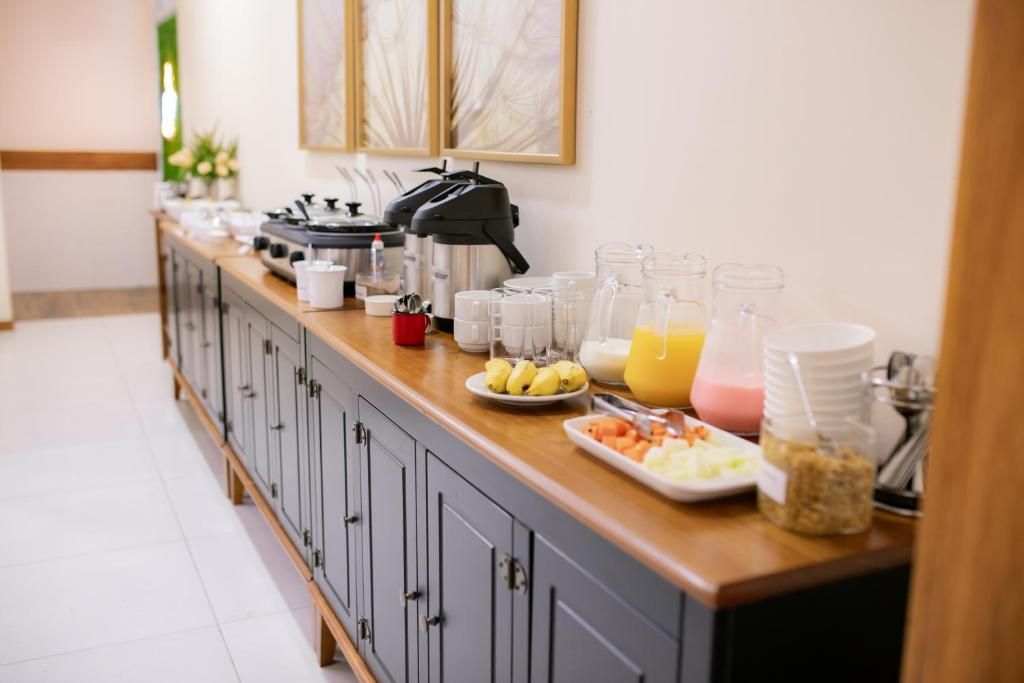 Aparador com cafeteiras, bebidas, frutas e cereais em um longo armário escuro contra uma parede clara.
