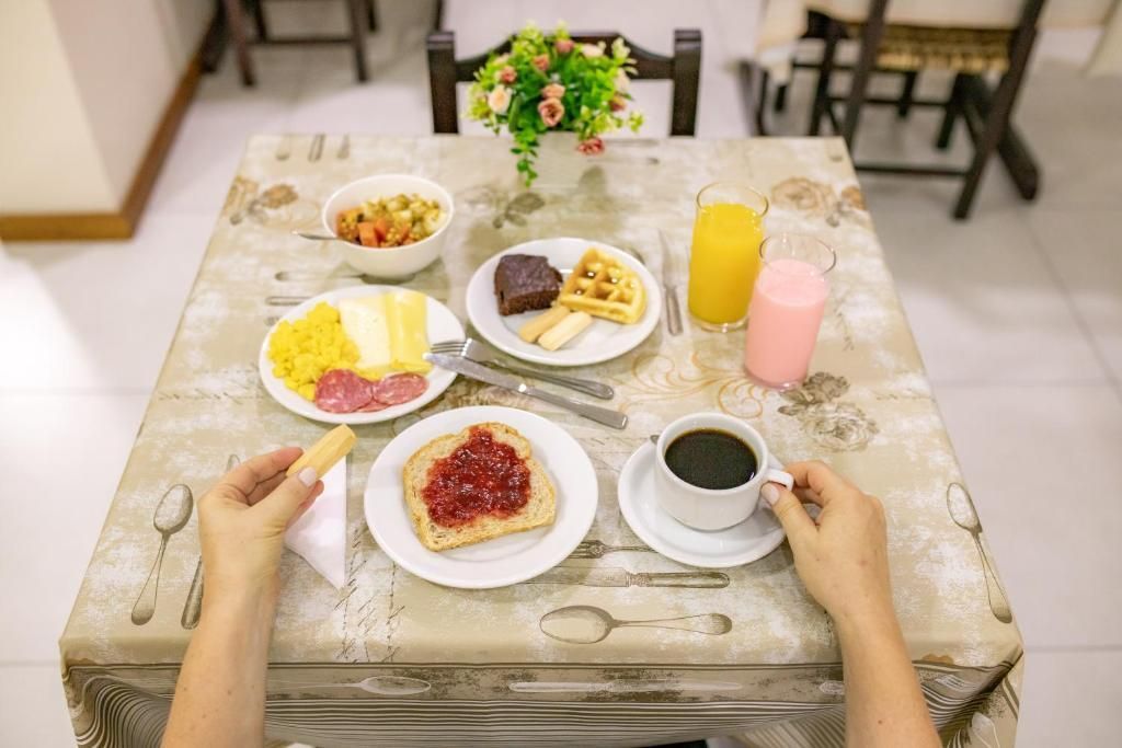 Mesa do café da manhã: torradas com geleia, waffles, frutas, ovos, café, suco, mãos segurando a comida.