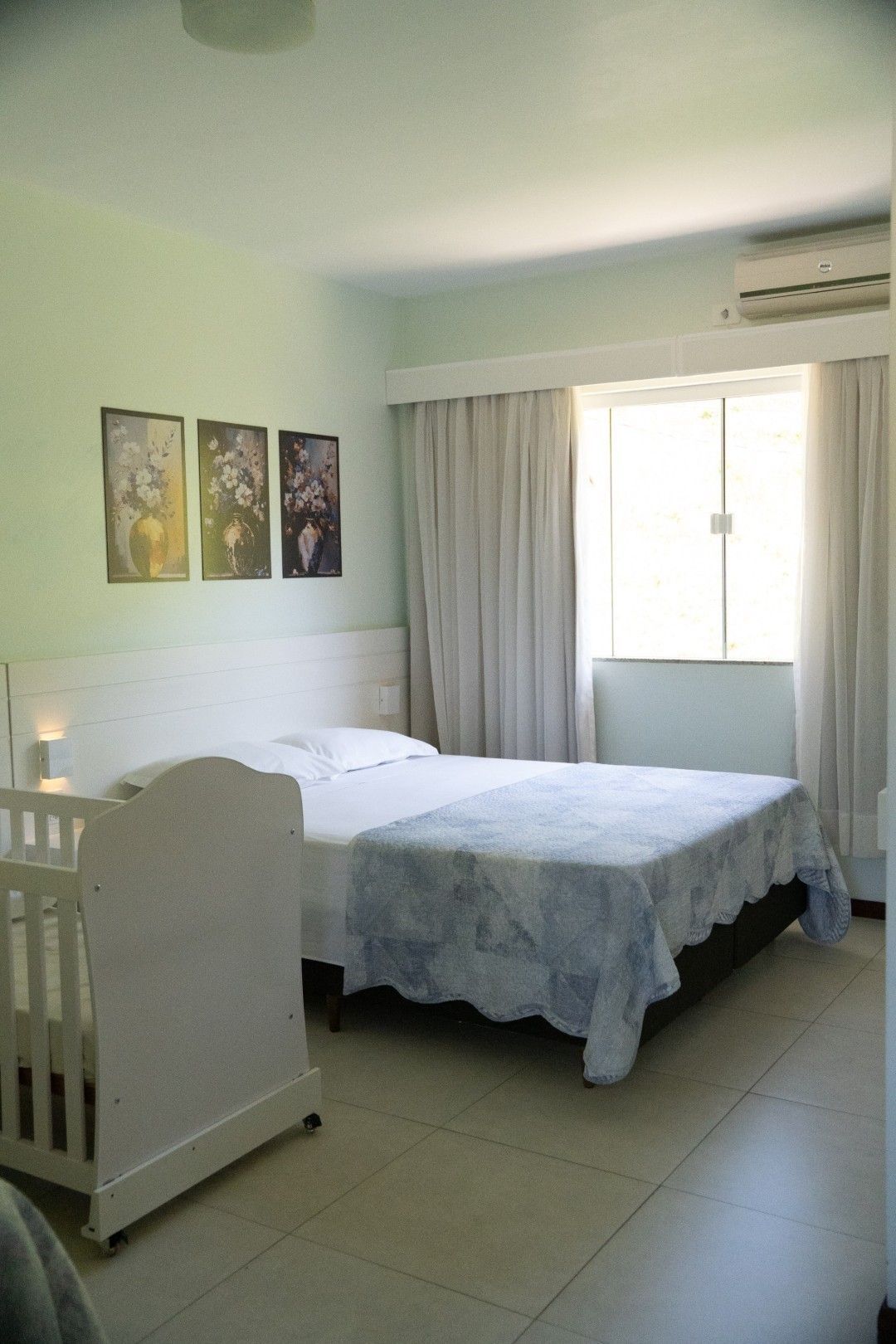 Quarto com cama, berço e janela com cortinas claras. Três obras de arte emolduradas estão penduradas acima da cama.