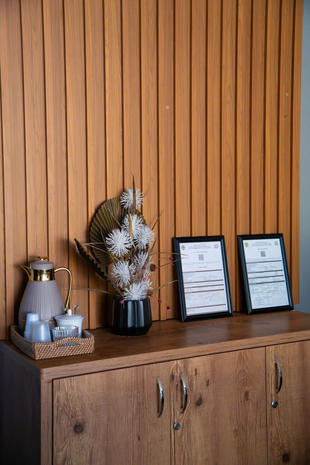 Armário de madeira com documentos emoldurados, arranjo de flores secas e bandeja com bule e xícaras de chá.