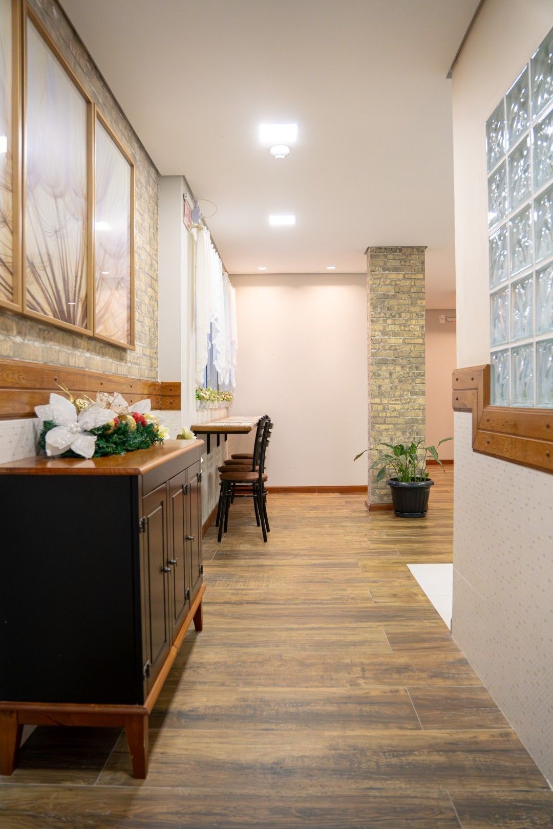 Corredor com piso de madeira, mesa lateral, mesa de jantar e pilar de pedra. Bem iluminado, com paredes em tons neutros e uma janela decorativa em blocos.