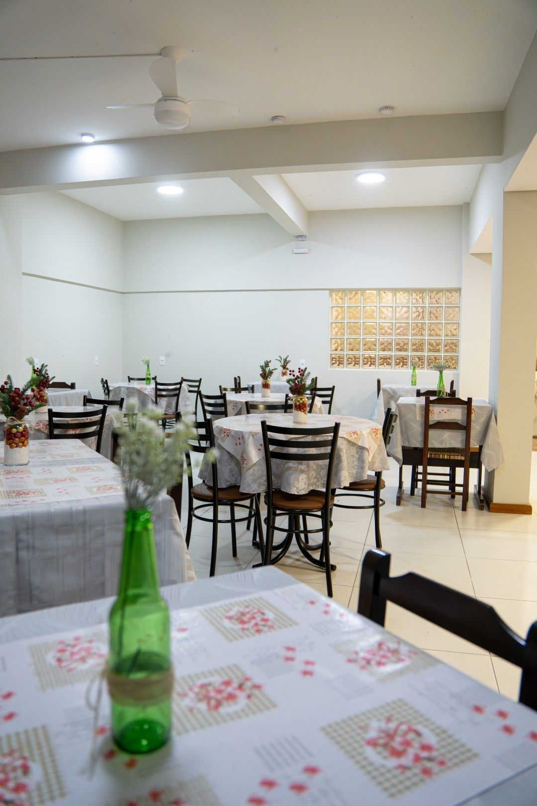 Sala de jantar vazia com mesas, cadeiras e arranjos de mesa sobre toalhas florais; uma garrafa verde em primeiro plano.