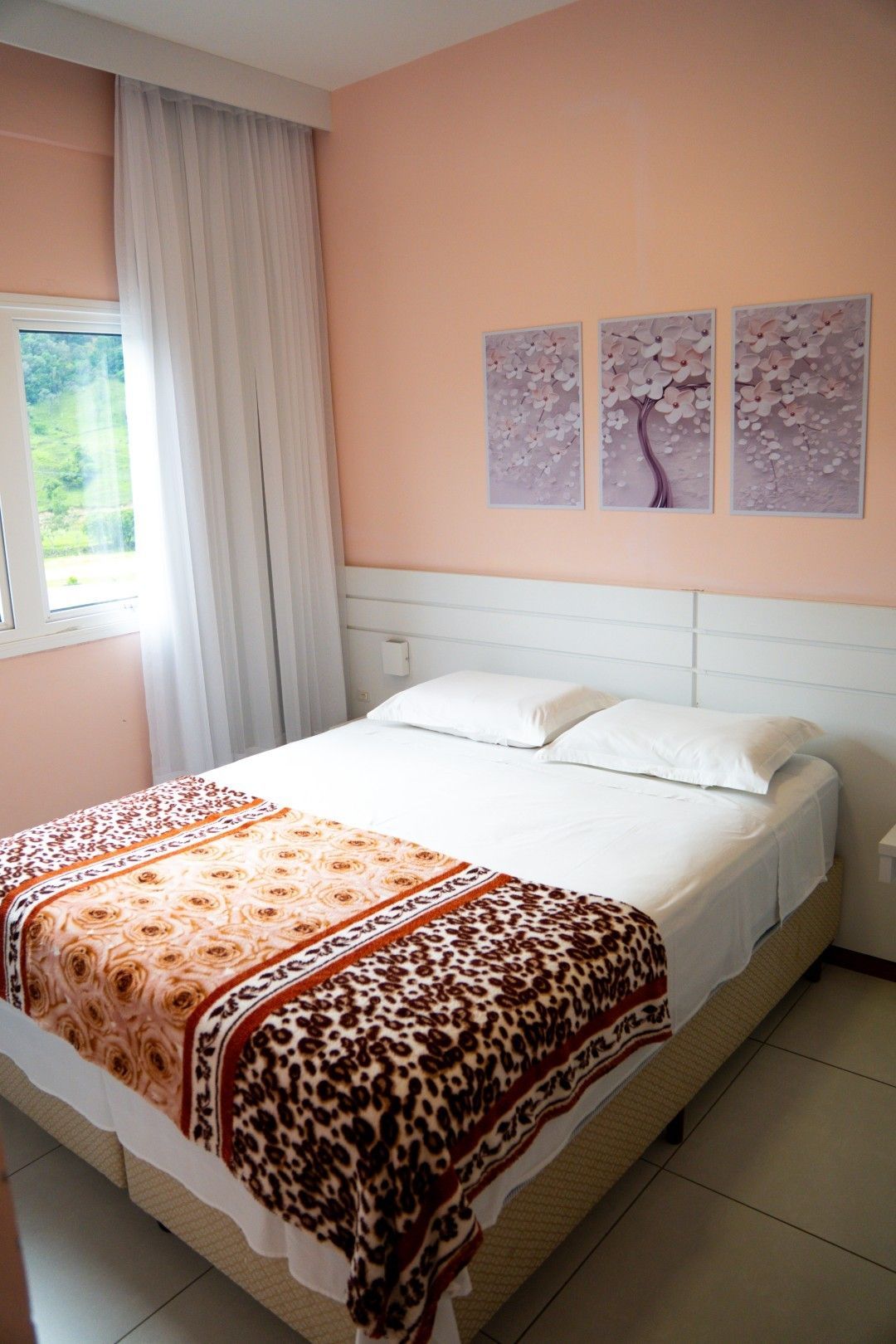 Quarto com cama de casal, paredes cor de pêssego, cortinas brancas e decoração floral na parede.