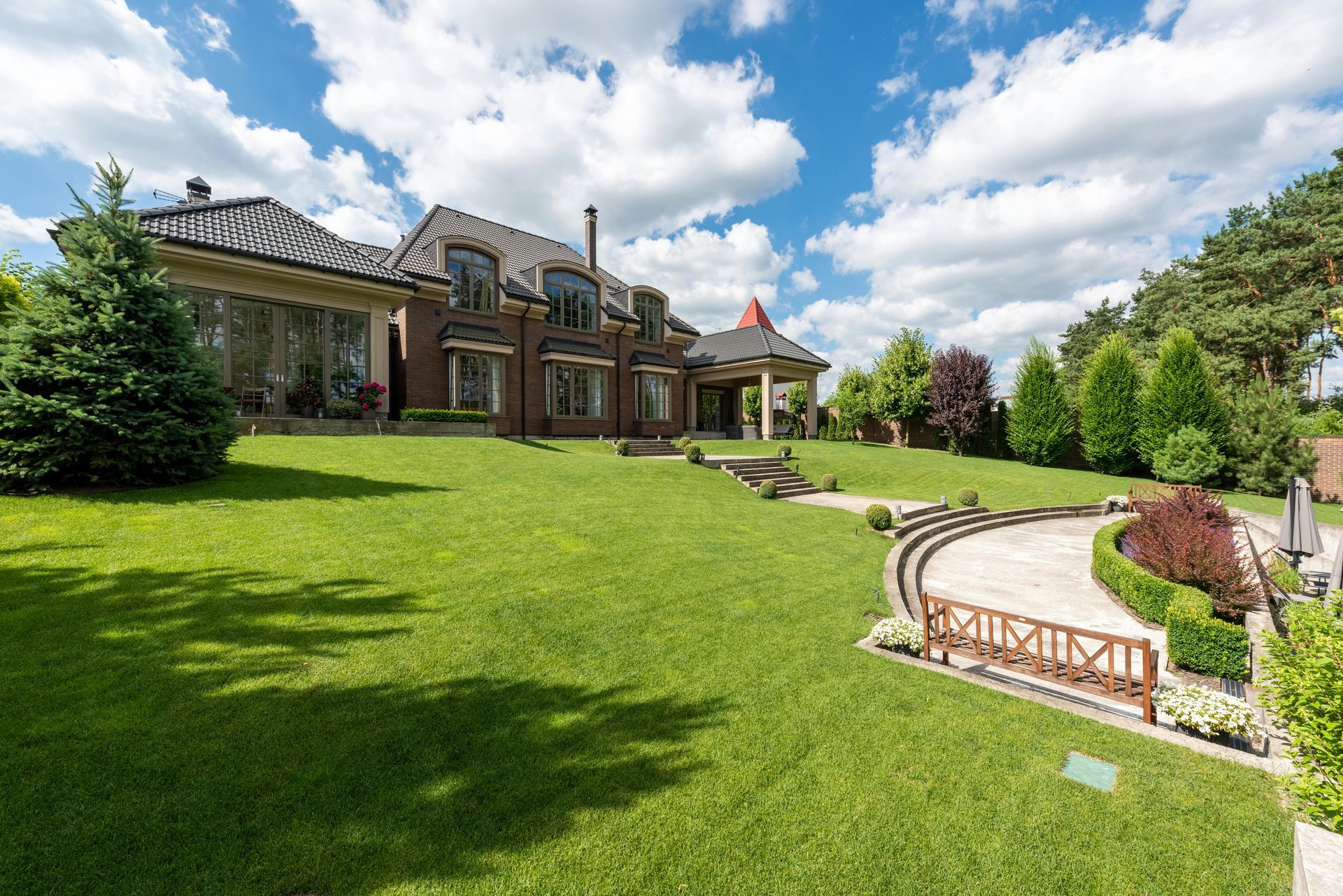 Large brick mansion with a lush green lawn, curved stone patio, and bright blue sky with clouds