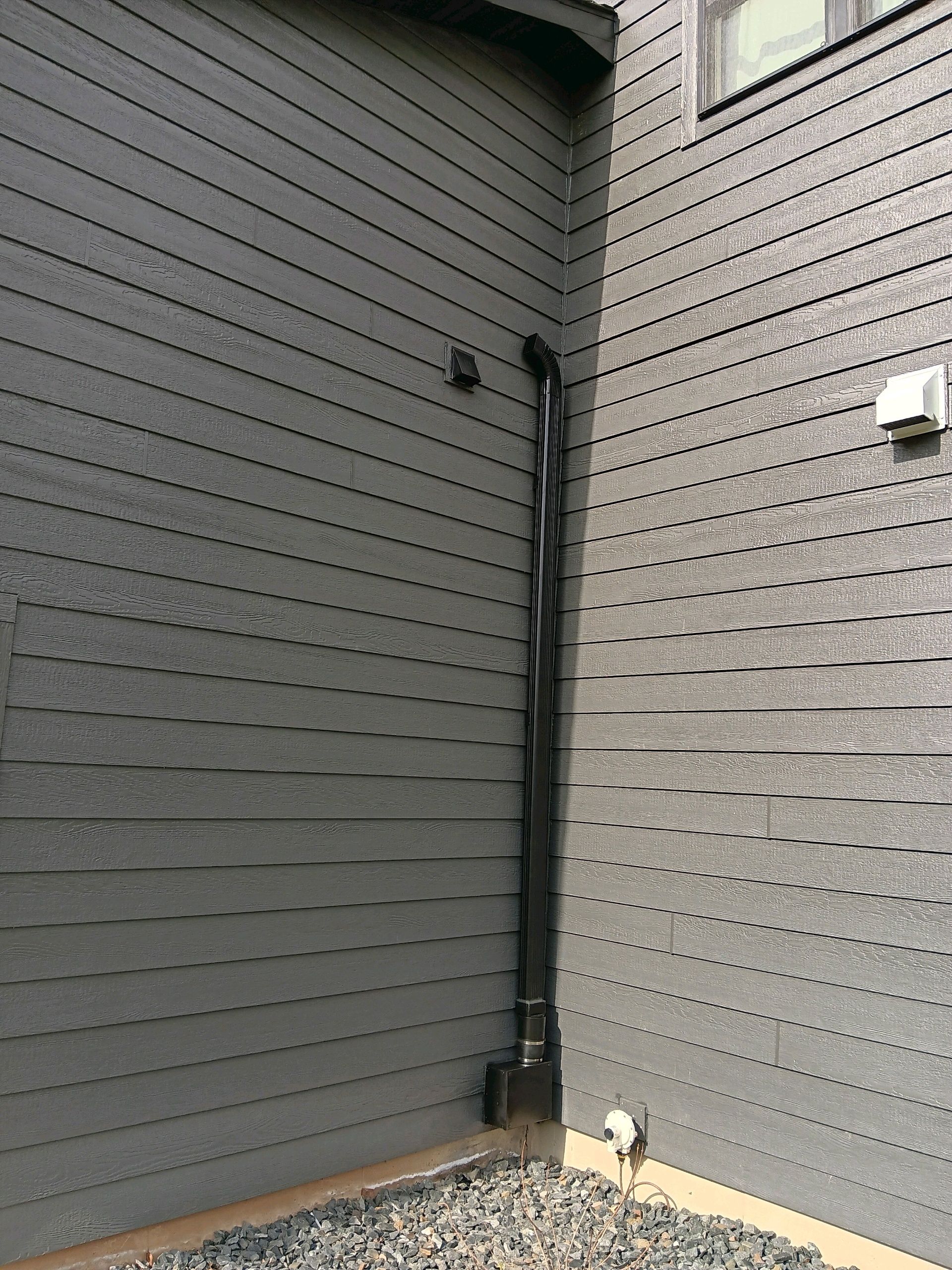 Black rain gutter running down a gray siding exterior wall to a gravel area.