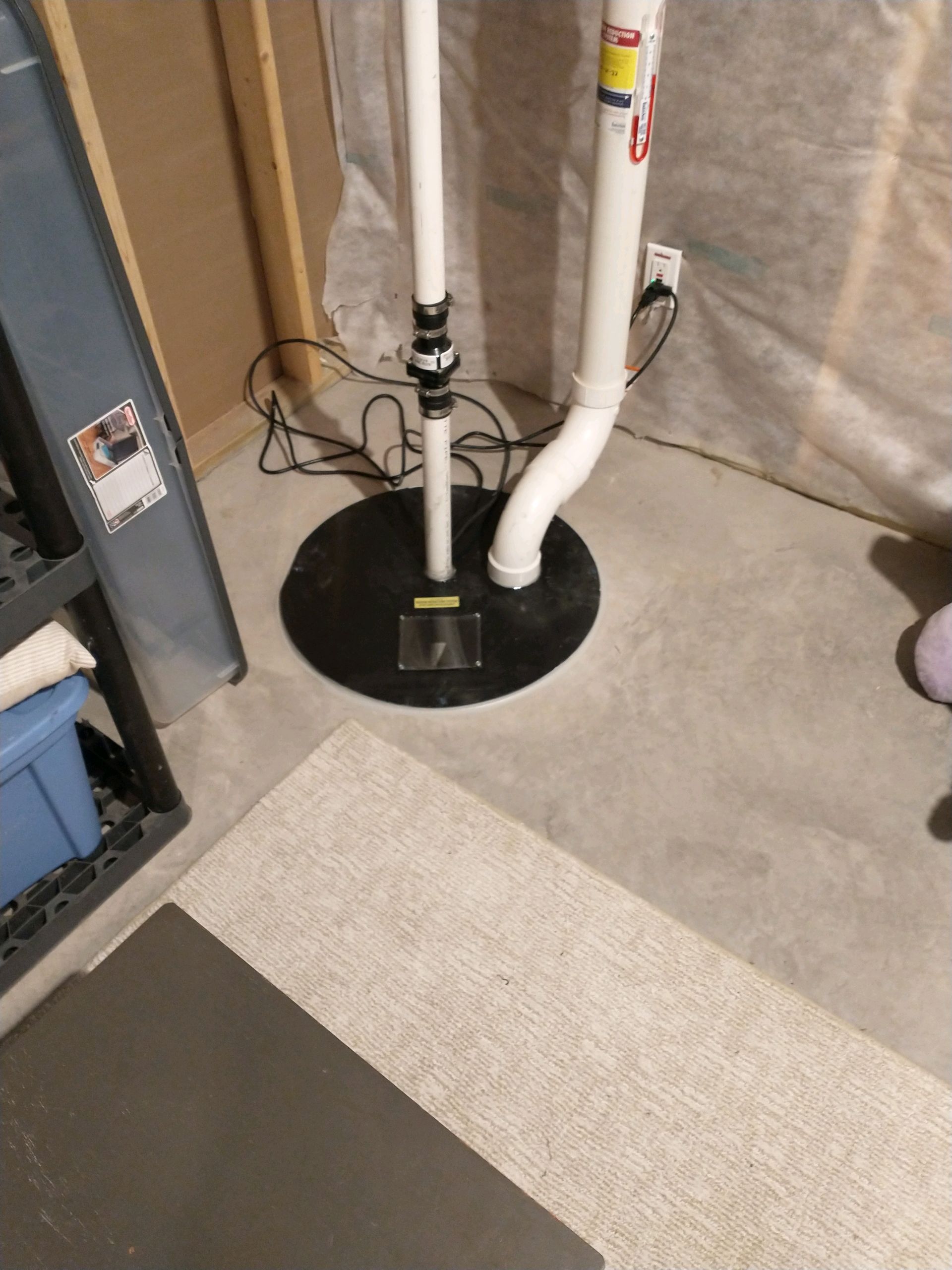 A sump pump with white pipes in a basement. The pump sits on a black base.