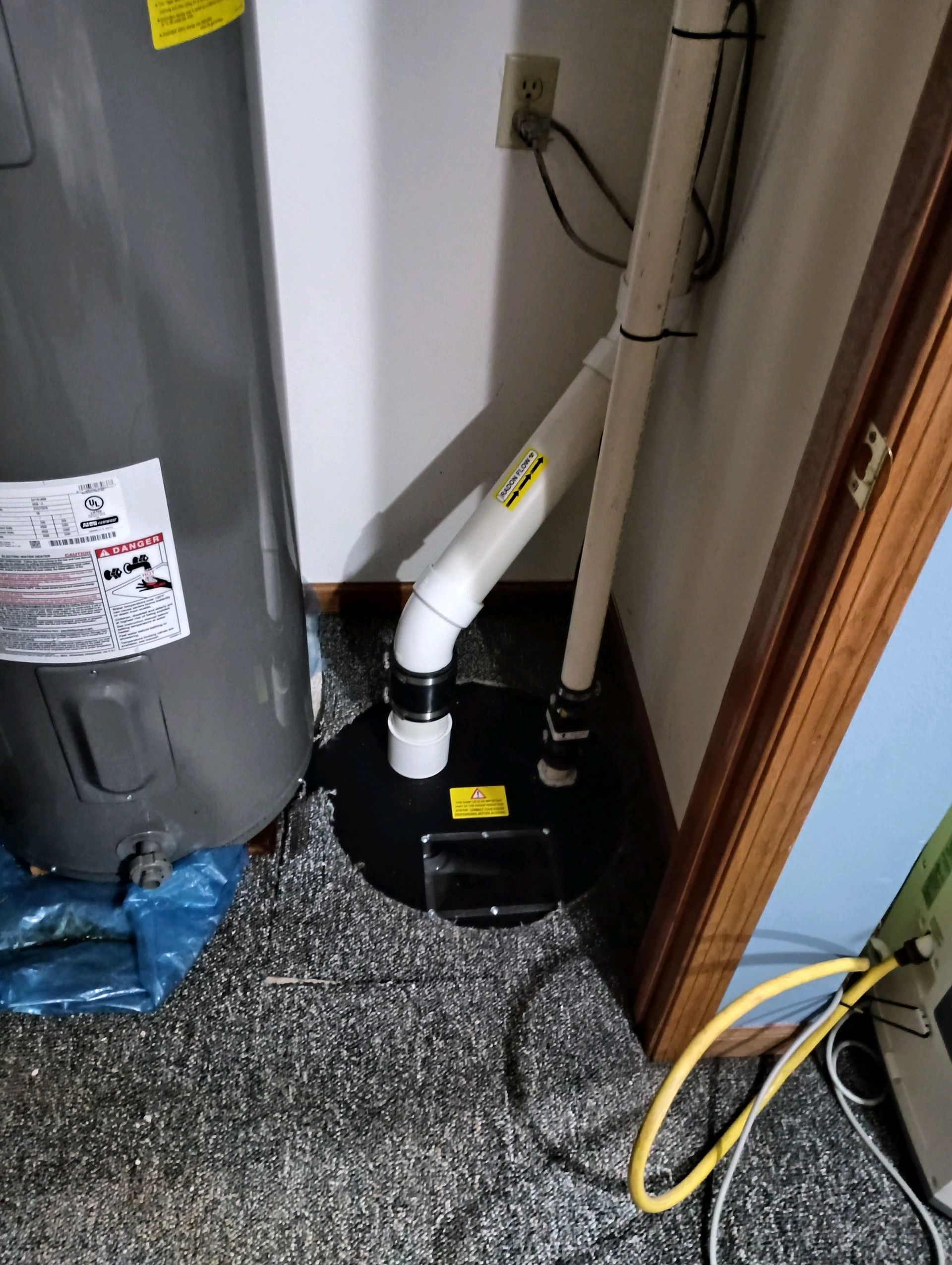 A sump pump in a basement corner, with white PVC pipes, near a water heater and a doorway.