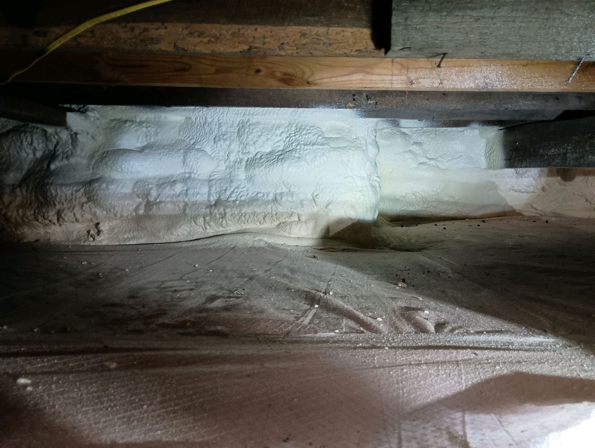 White spray foam insulation fills a crawlspace between wooden beams and floorboards.