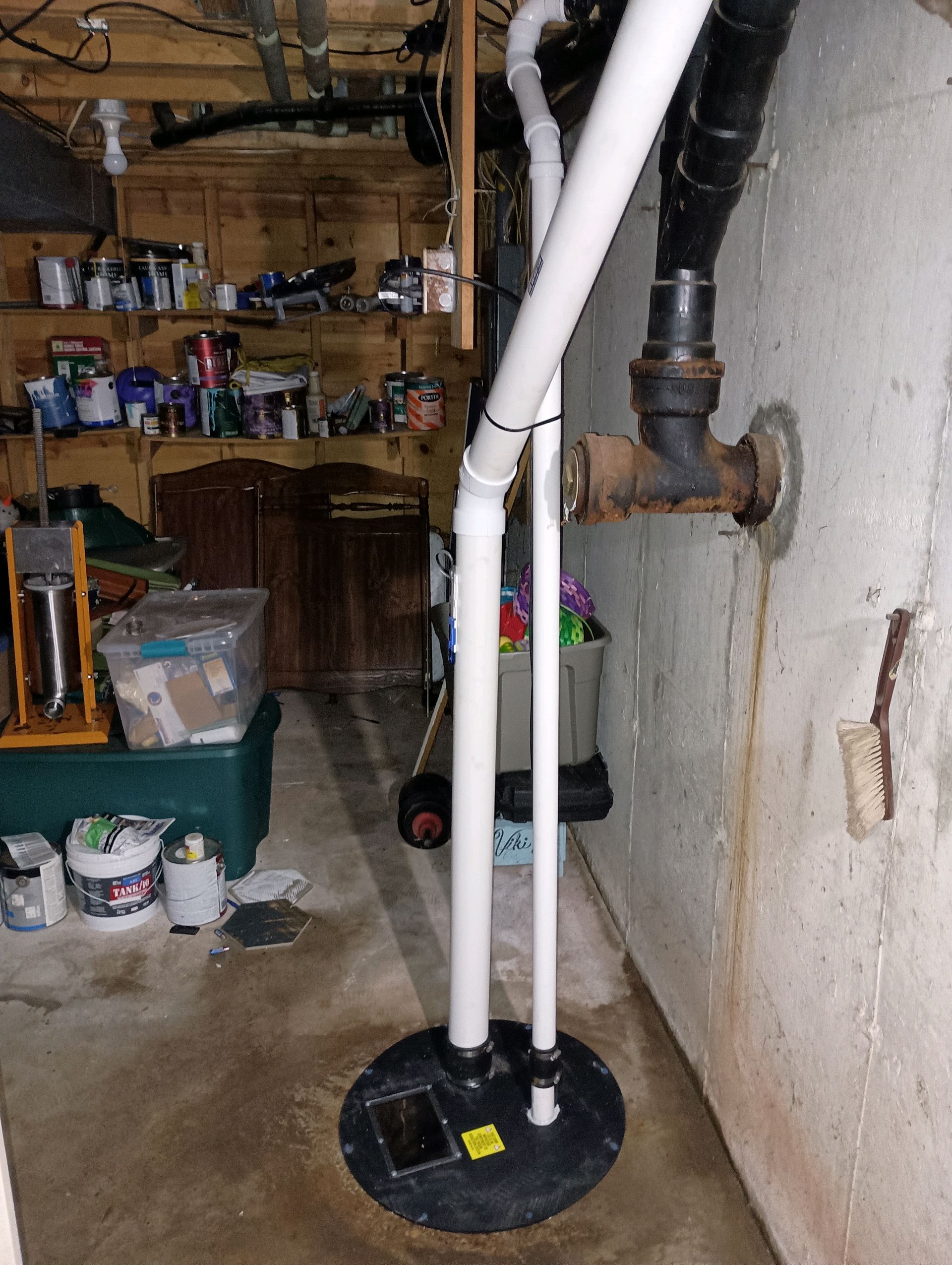 Basement with sump pump. Black, circular pump base with white PVC pipes against a concrete wall.