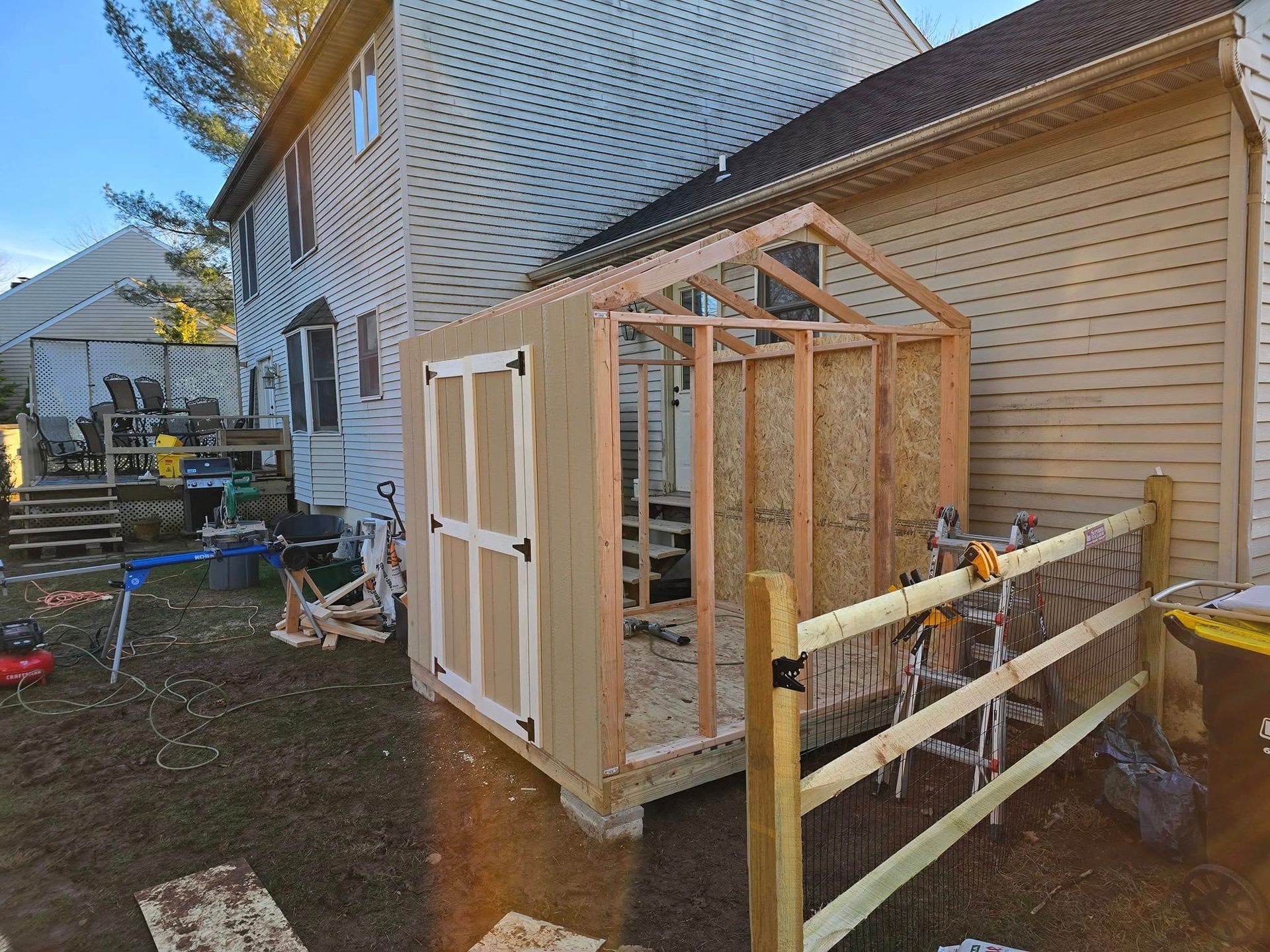 A partially constructed shed beside a two-story house, with wooden framing, door, and a wooden fence.