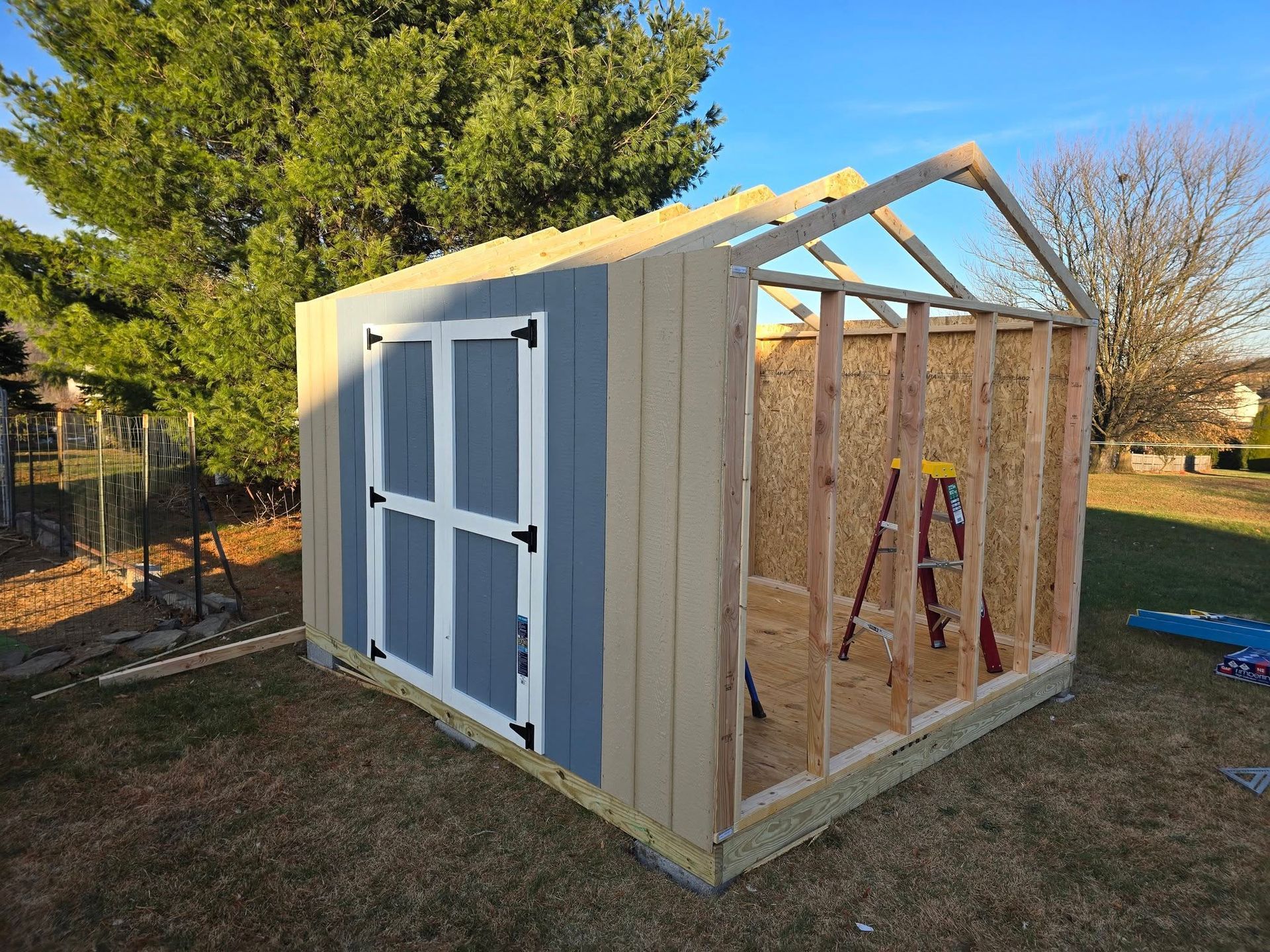 Partially built shed in a yard, with blue and tan siding, wooden frame, and door.