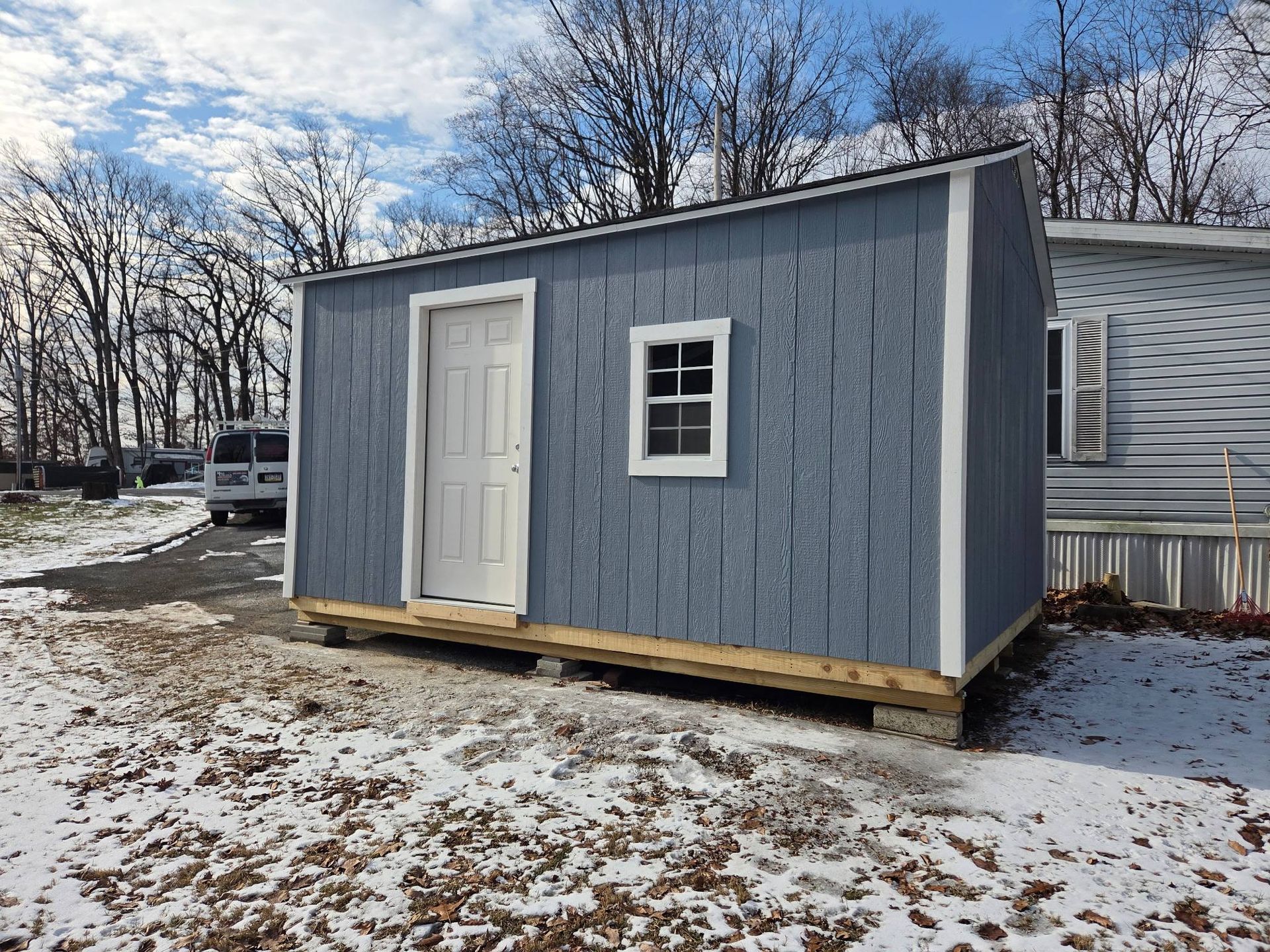 Blue shed with white door and window, set on wooden base, in snowy yard.
