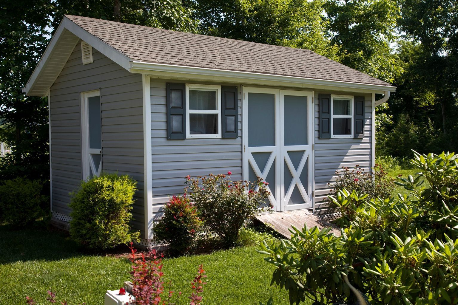 Gray shed with white trim, double doors, two windows, and dark roof in a grassy yard.