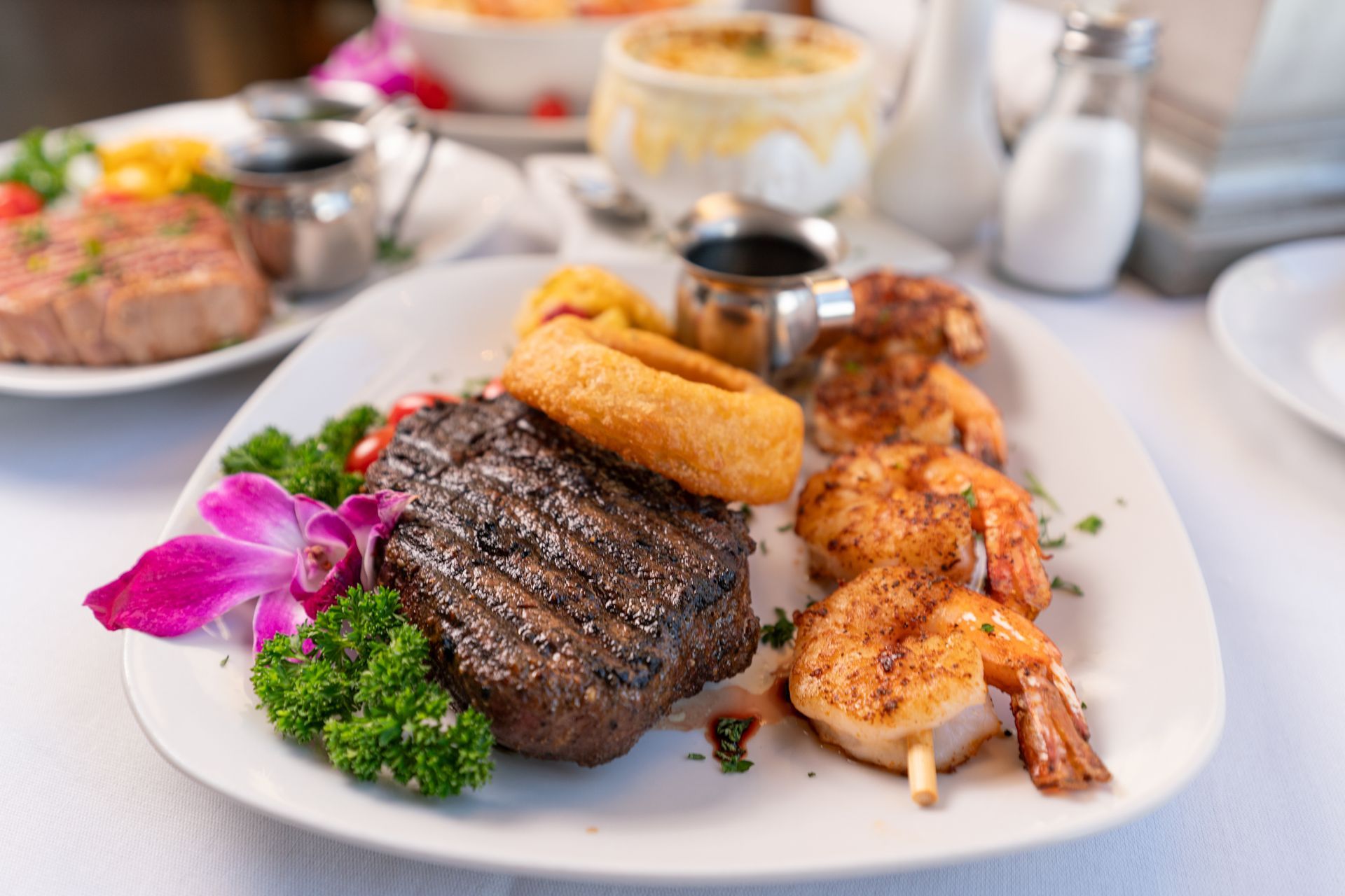 Steak dinner with shrimp and sides on a white plate, garnished with parsley and a purple orchid.