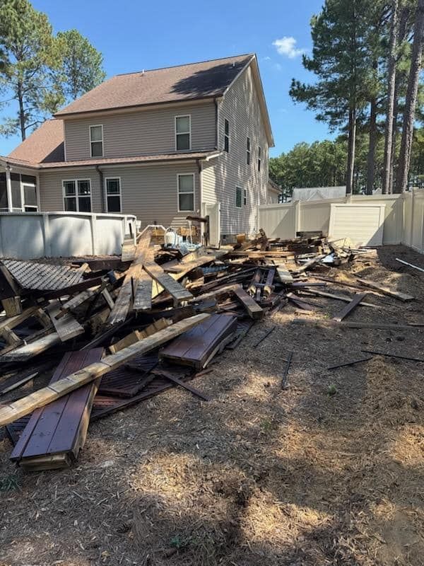 A pile of discarded wooden deck debris sits in a residential backyard next to an above-ground pool and beige house.