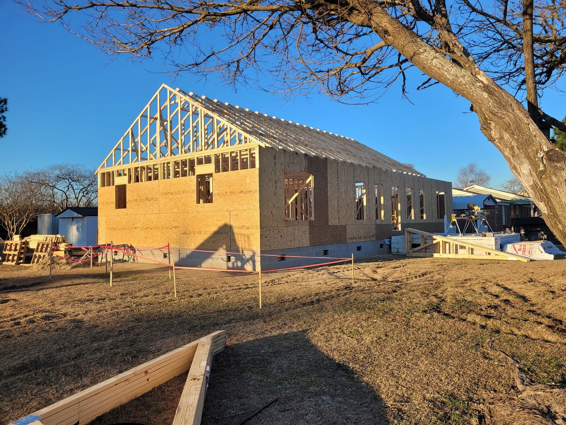 A newly framed wooden house under construction in an open, grassy lot on a clear, sunny day.